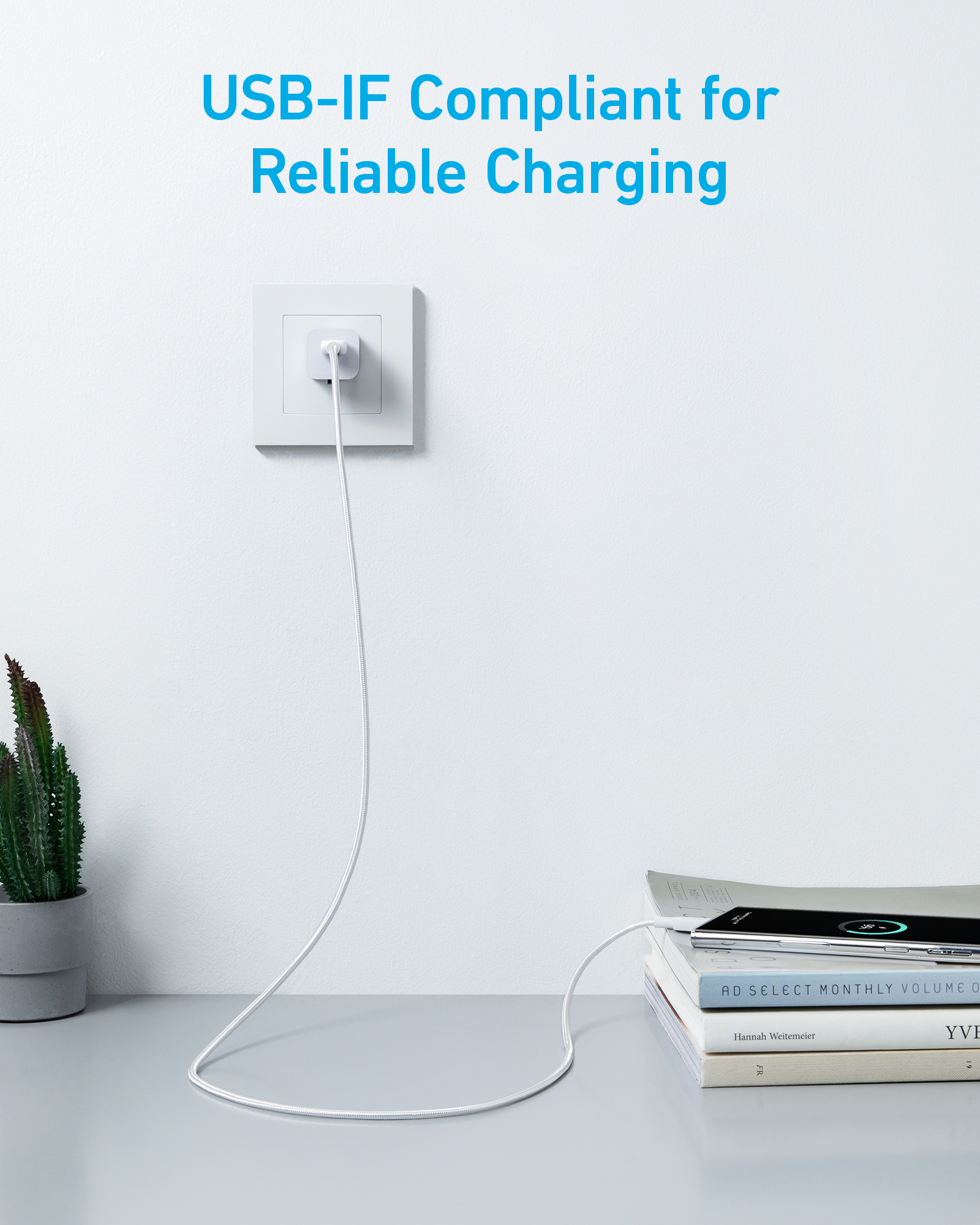 Un câble USB blanc charge un smartphone posé sur des livres. Un cactus est à proximité.