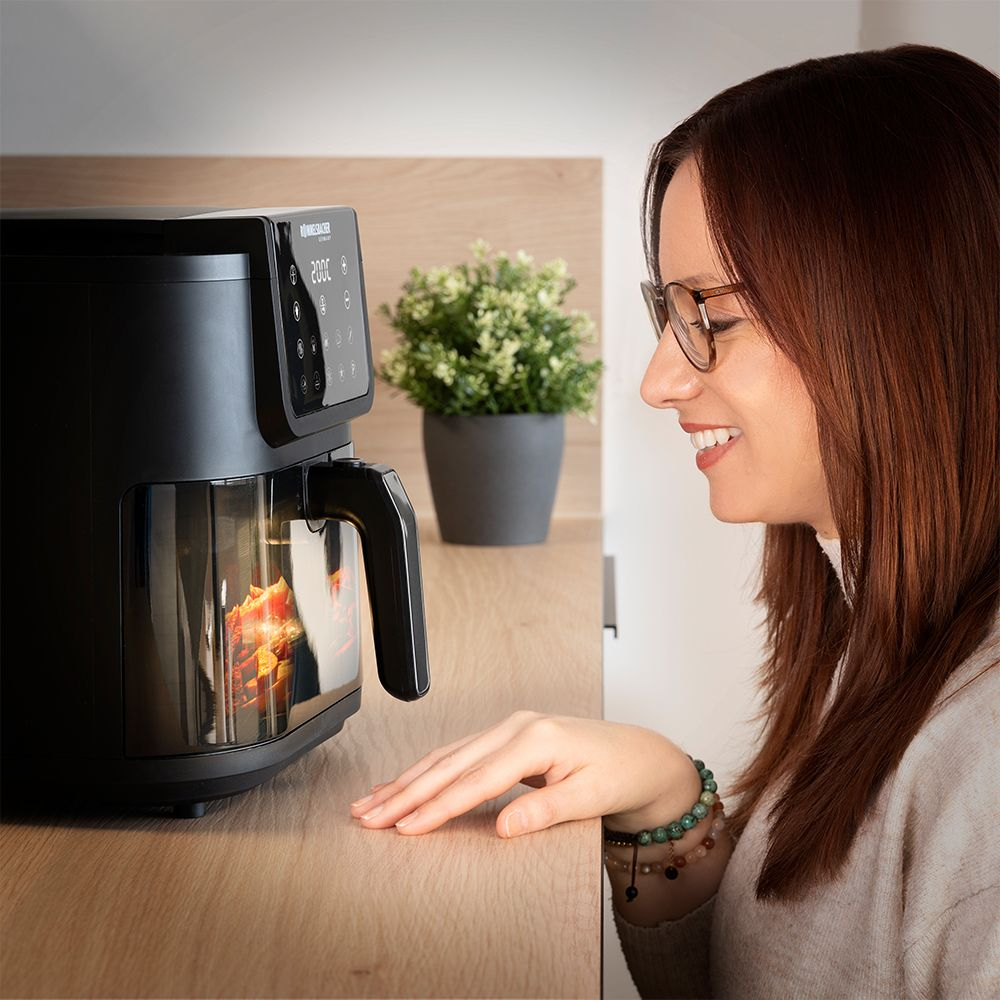 Kobieta patrzy na frytkownicę powietrzną gotującą jedzenie. Obok stoi zielona roślina w doniczce.