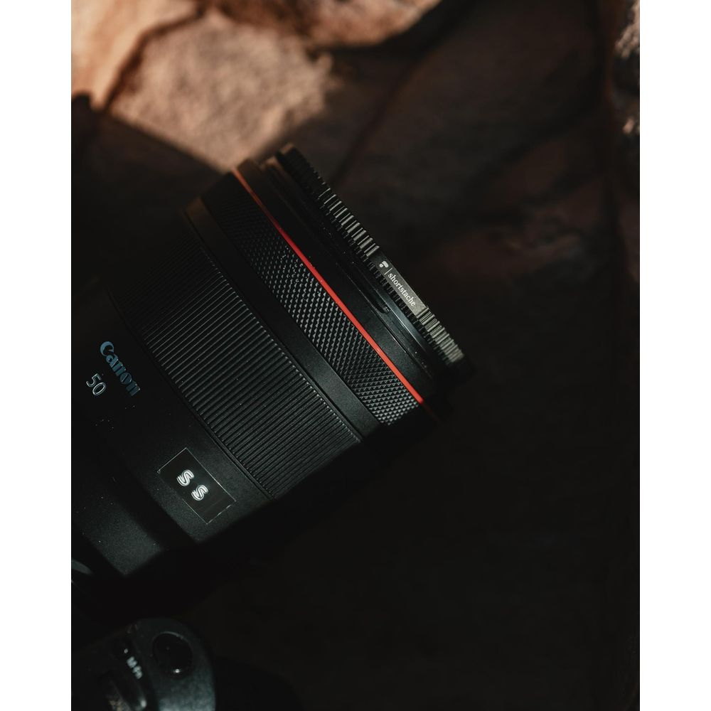A black Canon camera lens with a red ring rests on a dark background.