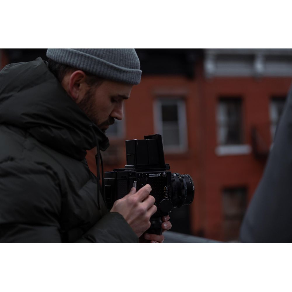 A man wearing a beanie and puffer jacket holds a camera in a street.