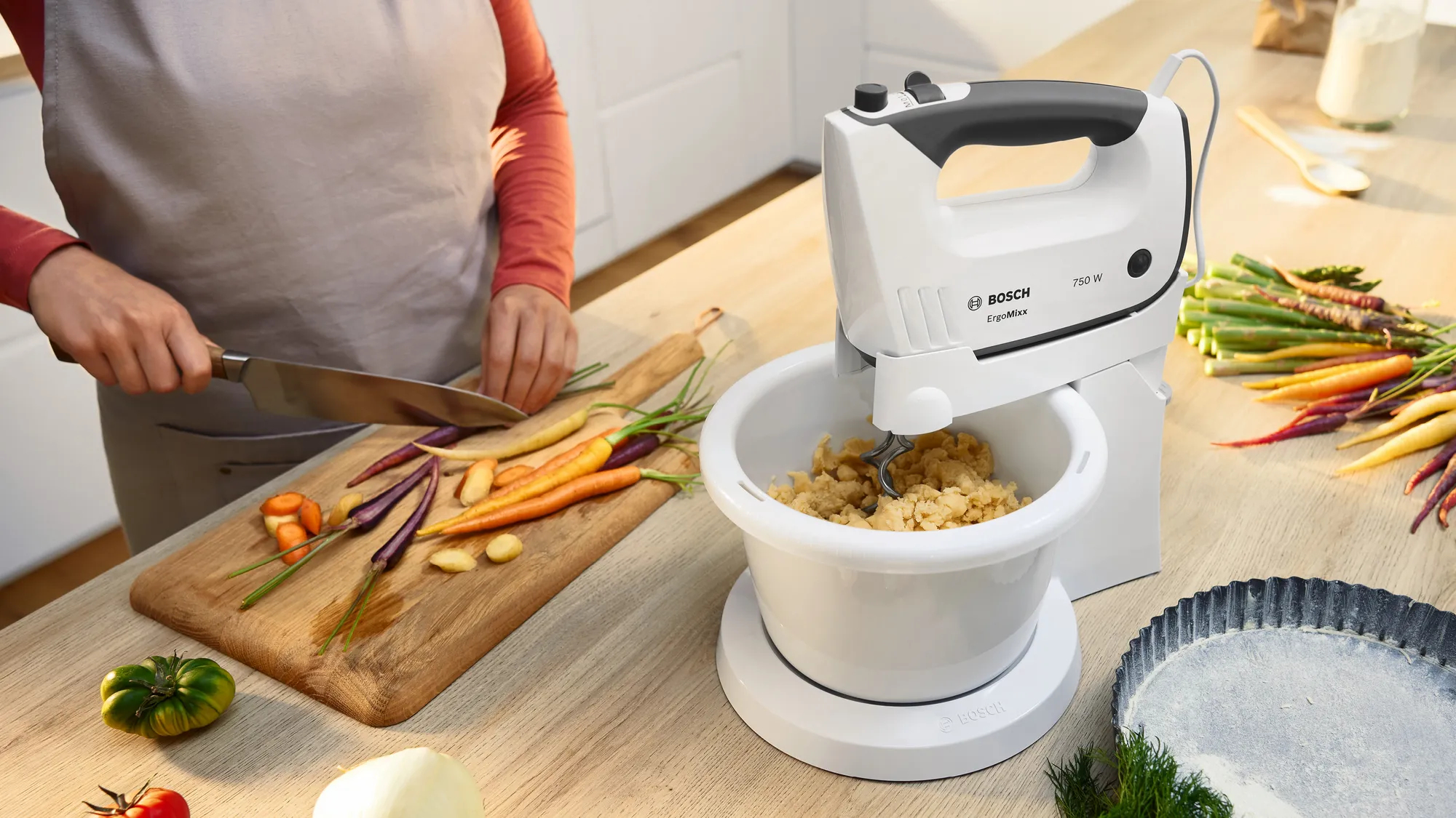 Eine Person schneidet Karotten, während ein Handmixer Teig in einer Schüssel mixt.