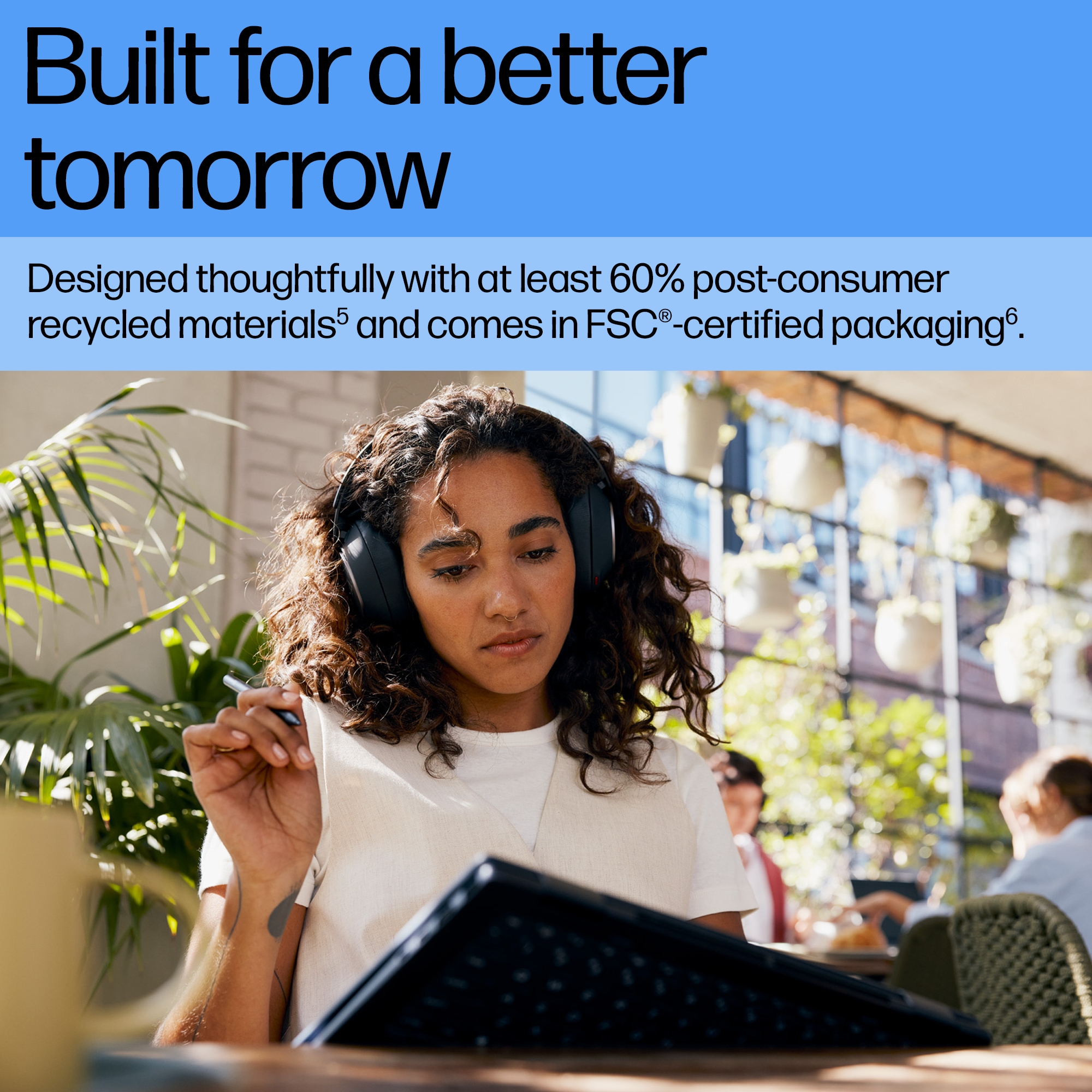 Kobieta w słuchawkach pracuje na laptopie na zewnątrz, z roślinami i ludźmi w tle.