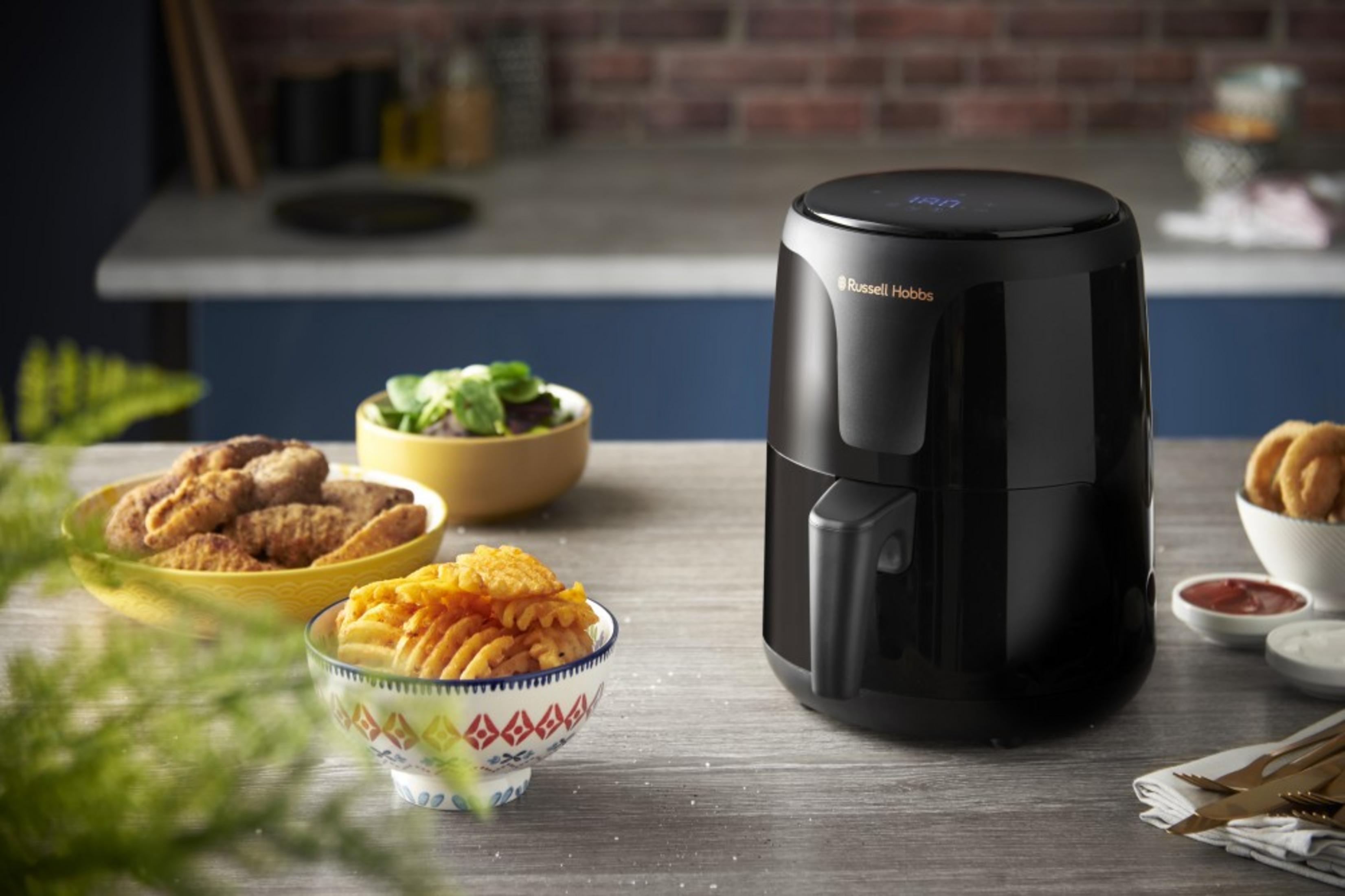 Una friggitrice ad aria Russell Hobbs nera accanto a ciotole di patatine, nugget e insalata.
