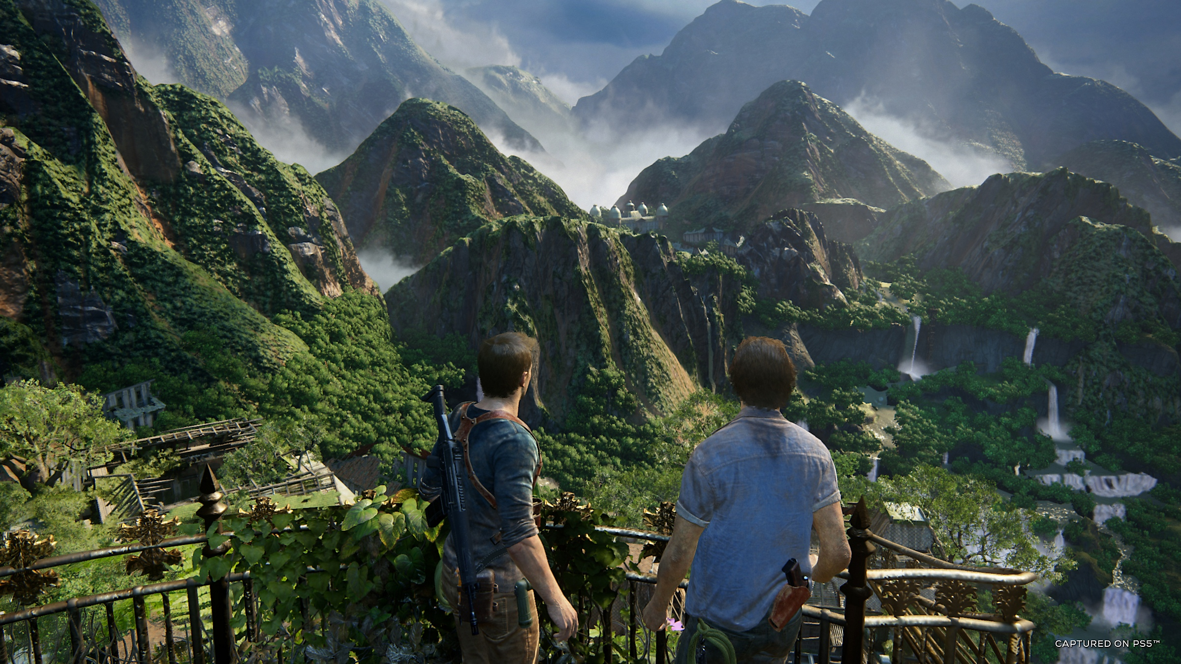 Two men stand on a balcony overlooking a lush mountain landscape with waterfalls.