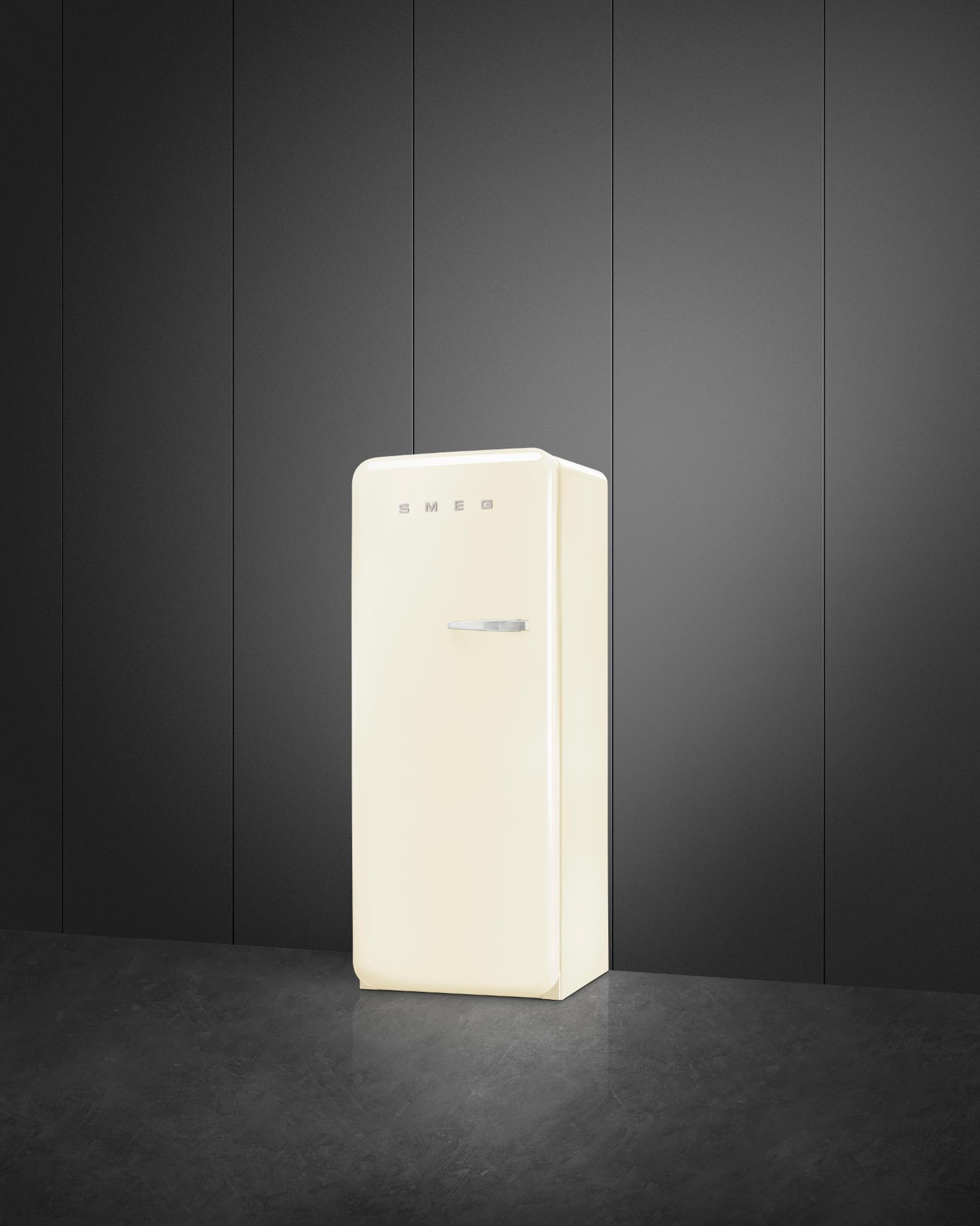 Ein cremefarbener Kühlschrank mit silbernem Griff steht auf dunklem Boden vor schwarzer Wand.