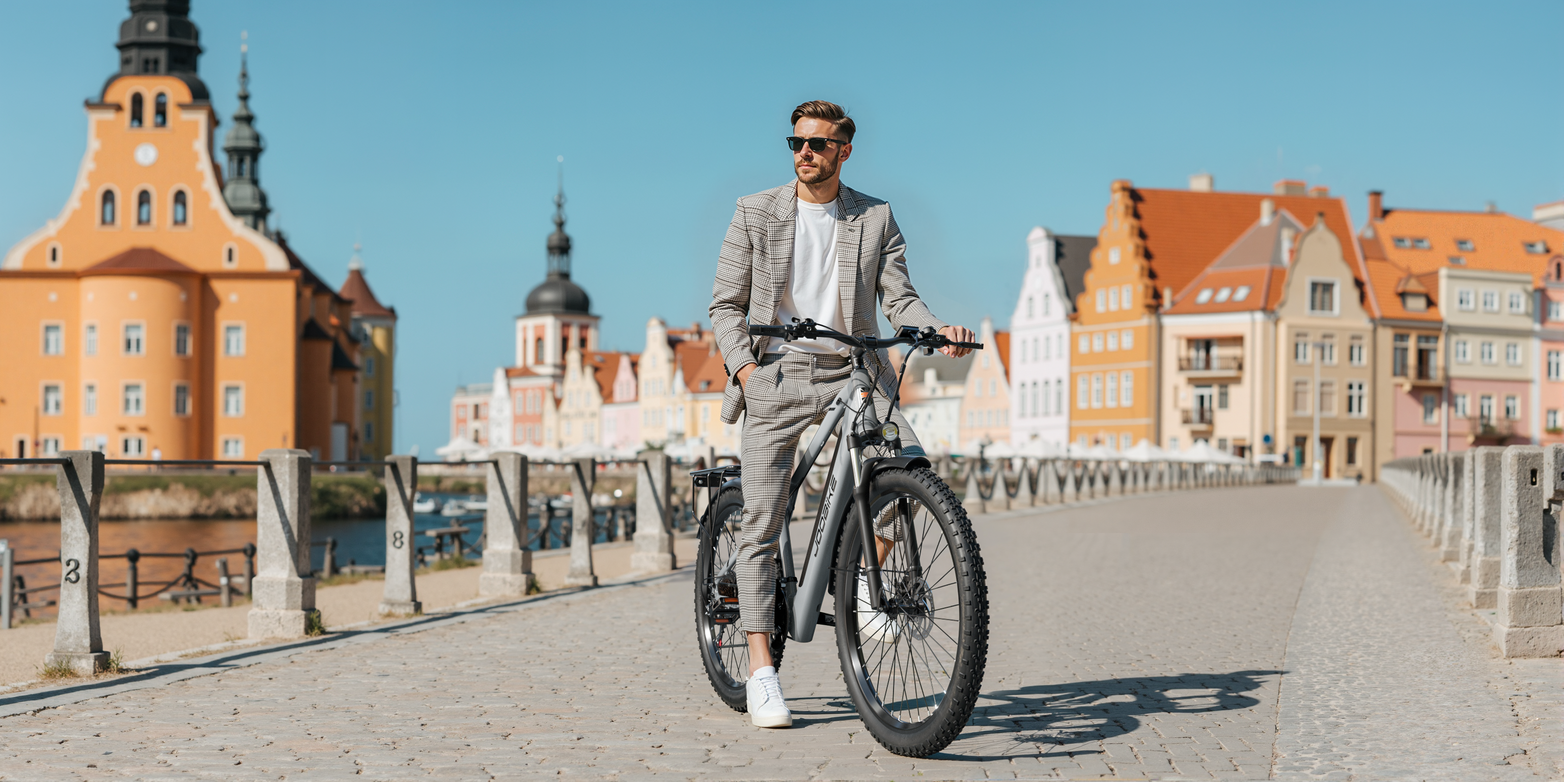 Mann im Anzug mit Fahrrad auf einer gepflasterten Straße vor bunten Gebäuden.