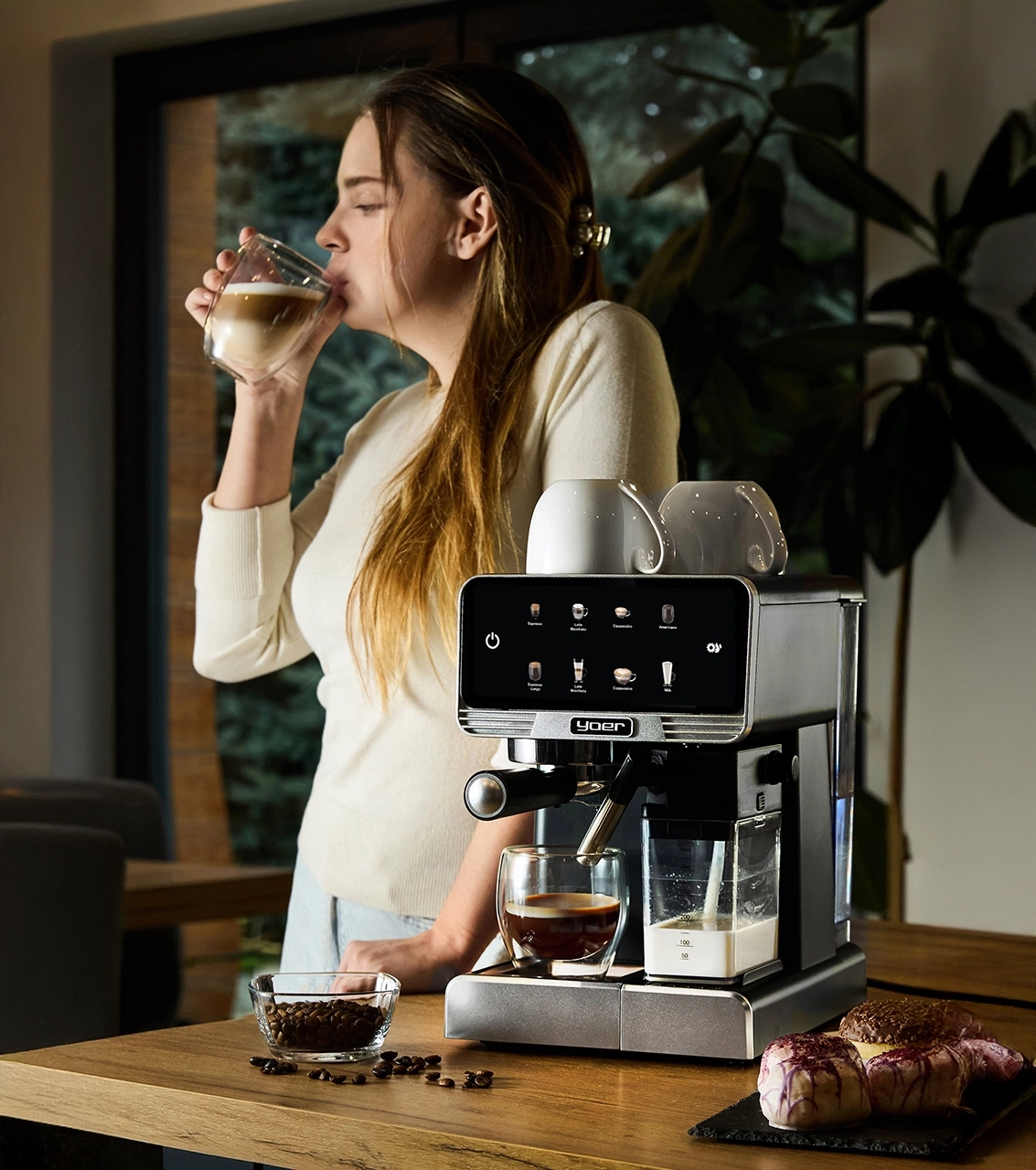 Kobieta pije kawę obok ekspresu do kawy ze szklanką mleka i pączkami.