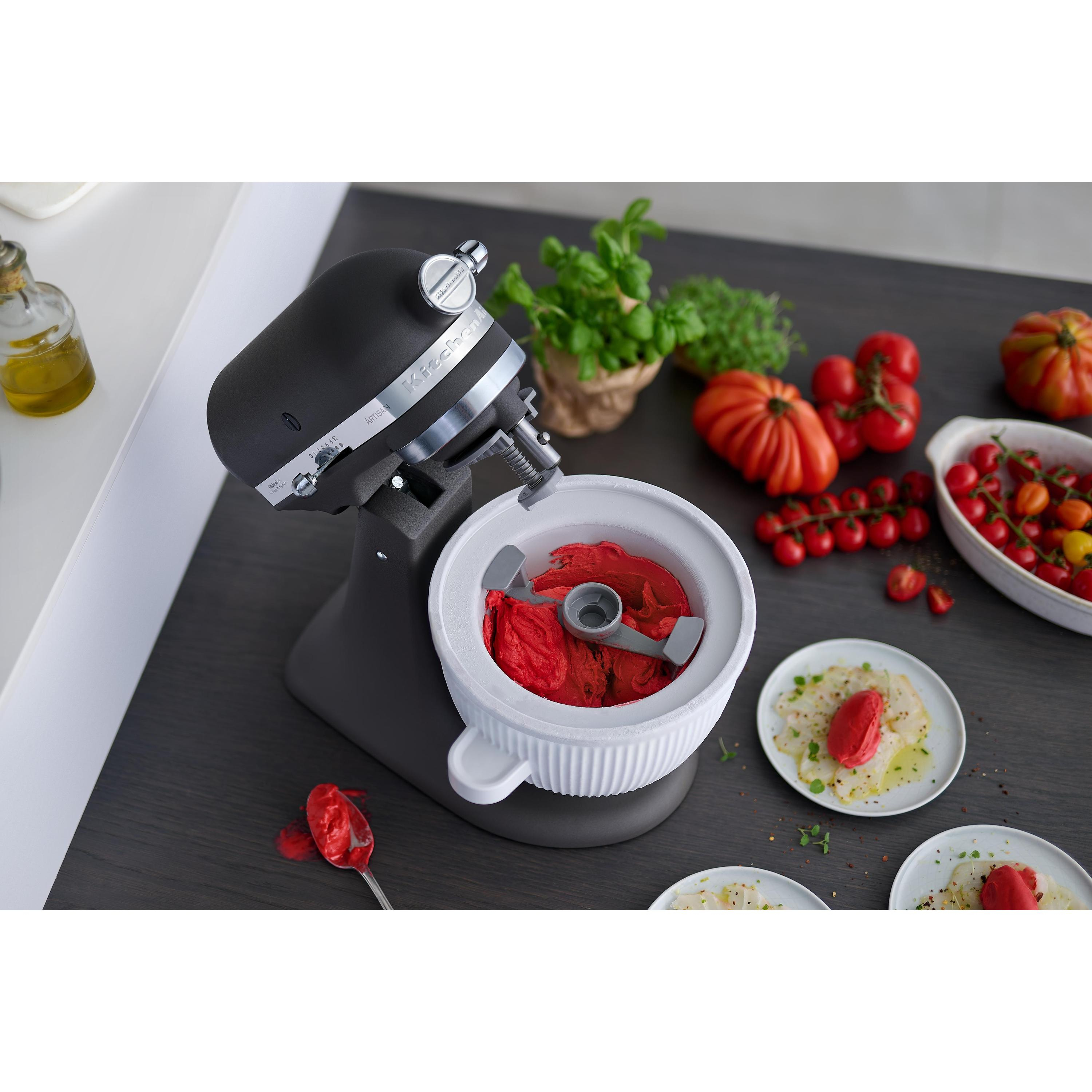 Ein schwarzer Küchenmaschinenaufsatz mit roter Masse in einer weißen Schüssel, daneben Tomaten.