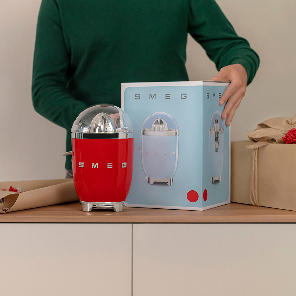 Osoba w zielonym swetrze rozpakowuje czerwony wyciskacz do cytrusów Smeg obok pudełka z produktem.