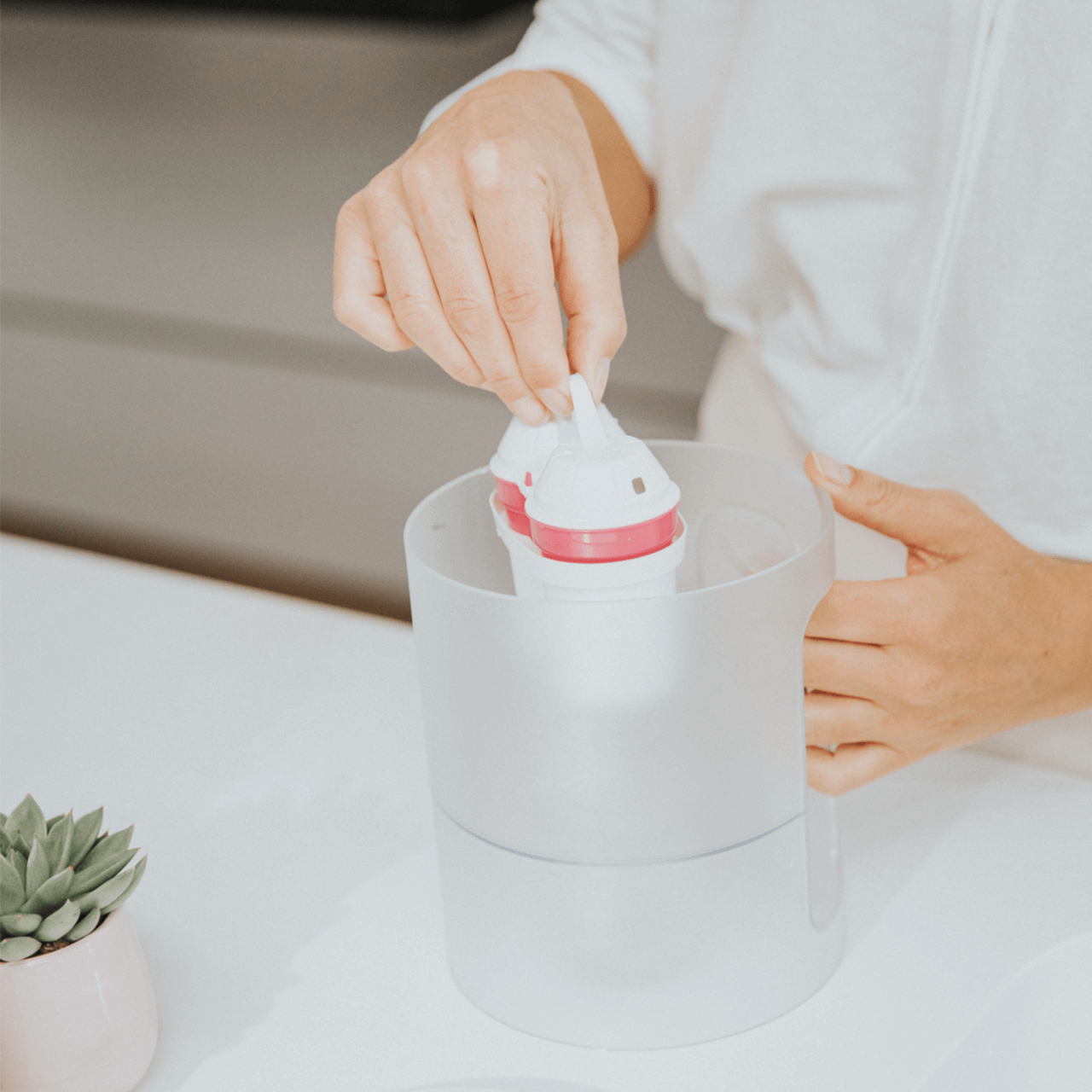 Person legt weiße Babyflaschen mit rosa Akzenten in einen transparenten Behälter.
