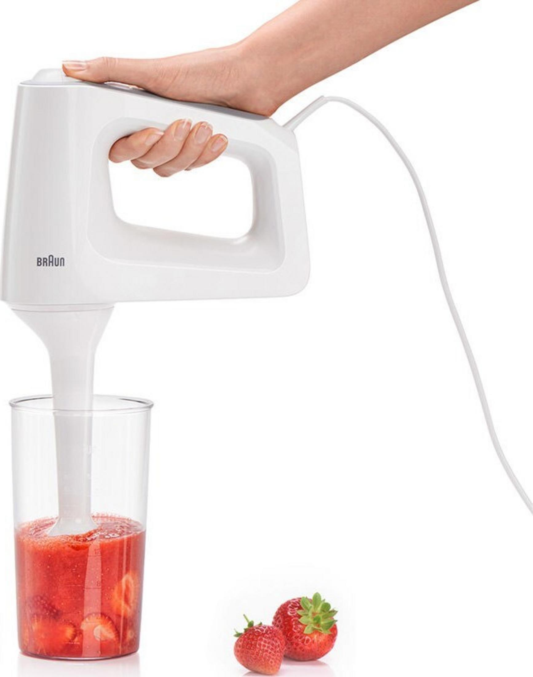 Een hand houdt een witte staafmixer die aardbeien in een glas mixt.