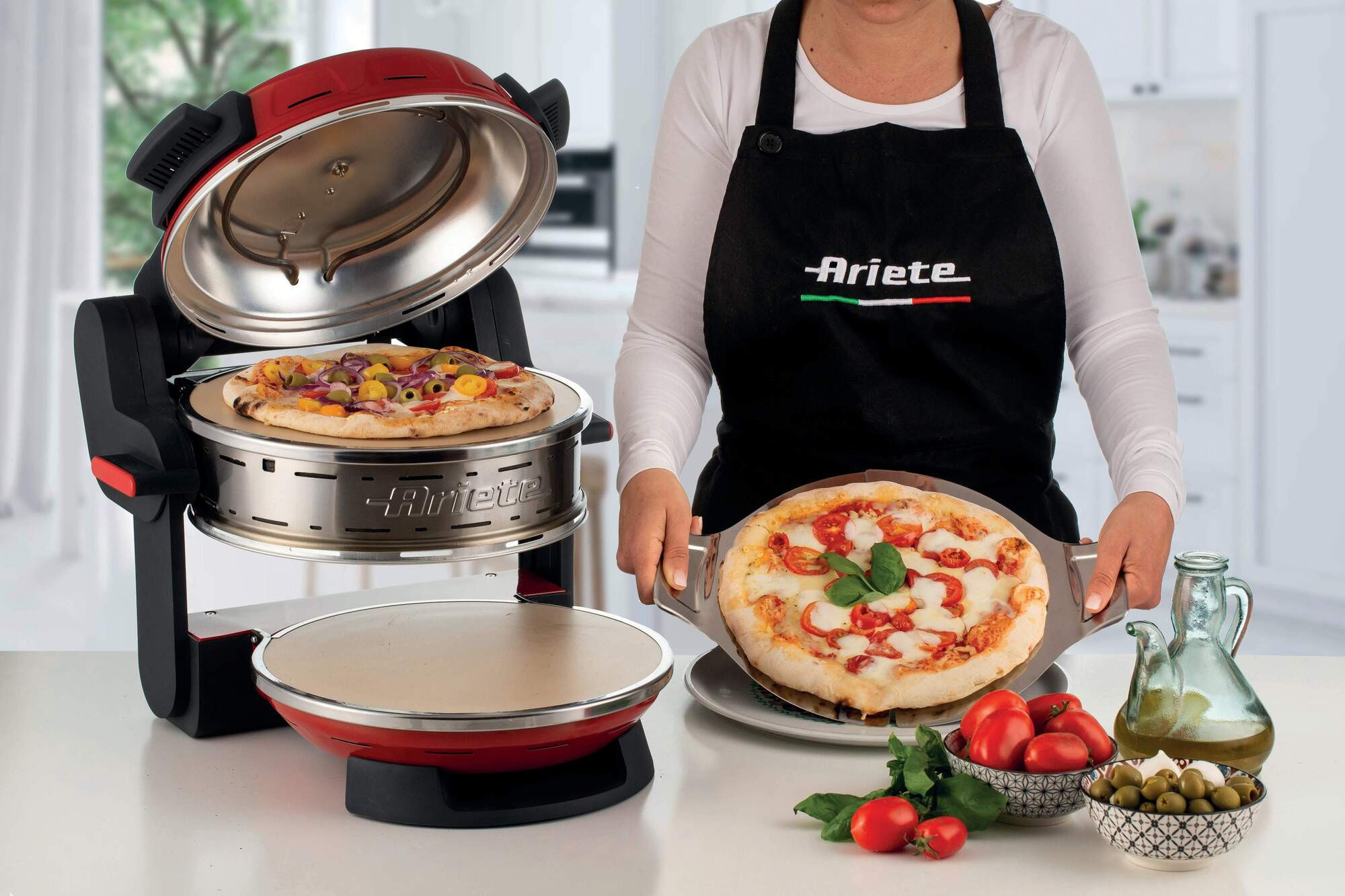 Eine Person in schwarzer Schürze hält eine Pizza. Ein Pizzaofen mit Pizza steht daneben.