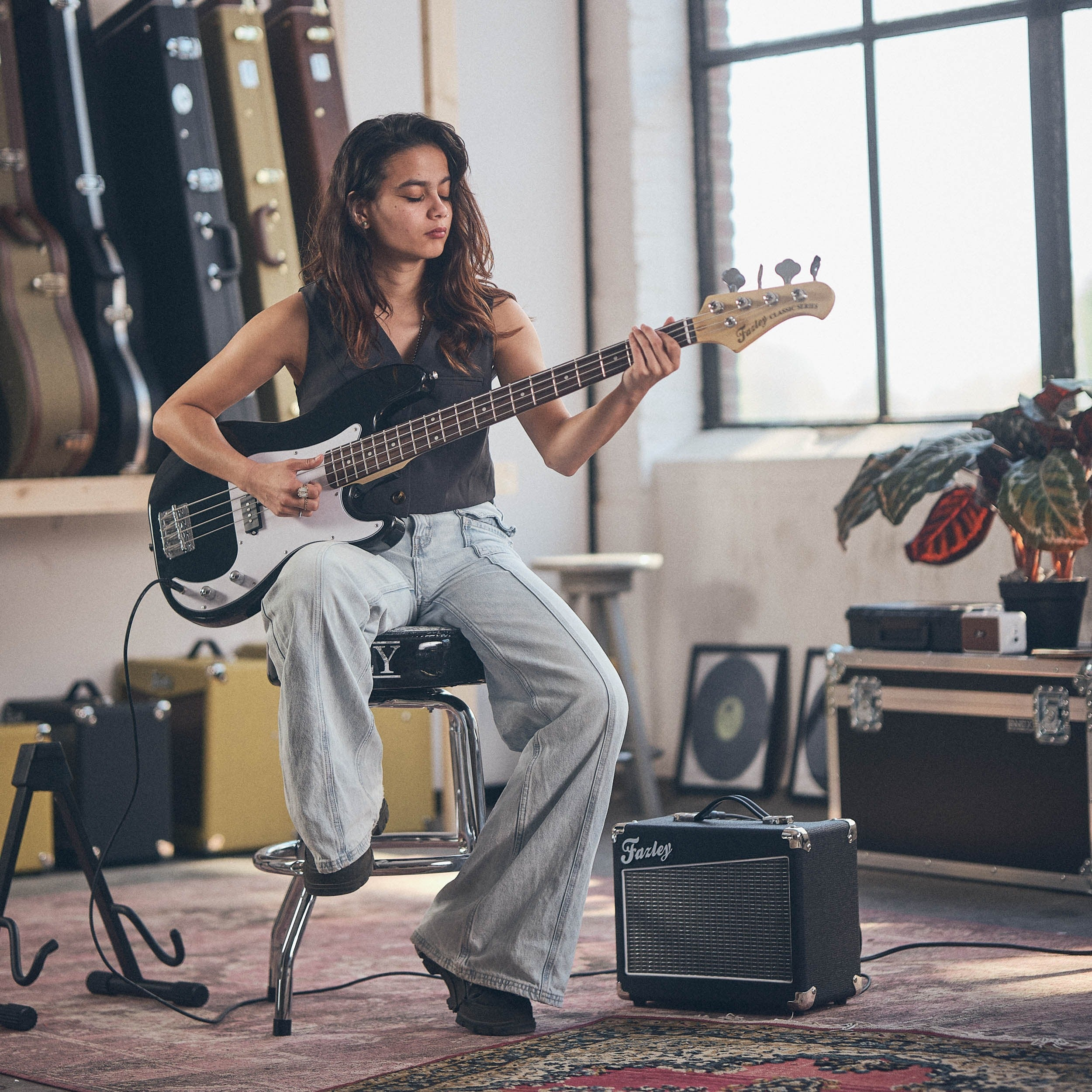 Vrouw speelt basgitaar in een kamer met versterkers en gitaarkoffers.