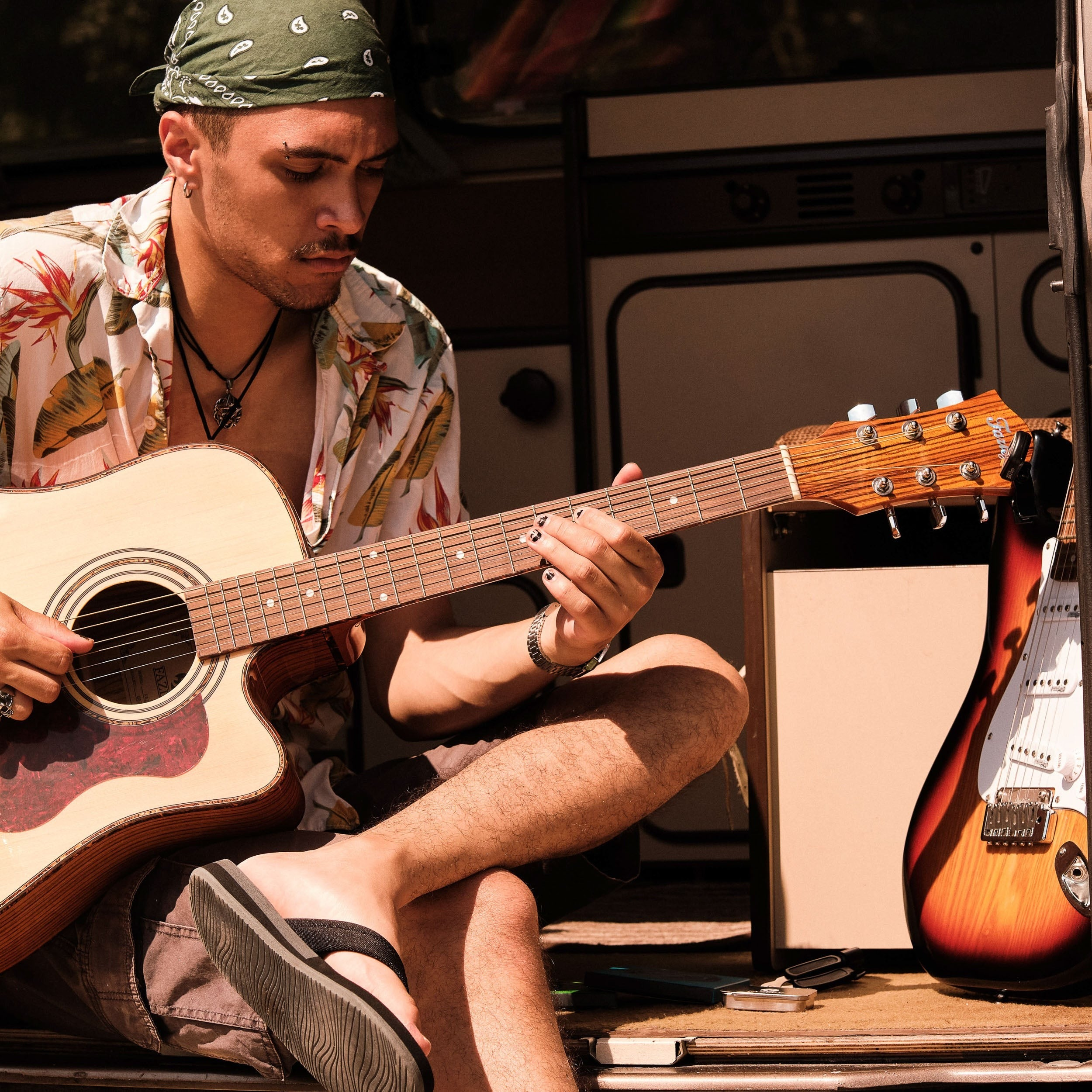 Man with bandana plays acoustic guitar, electric guitar beside him.