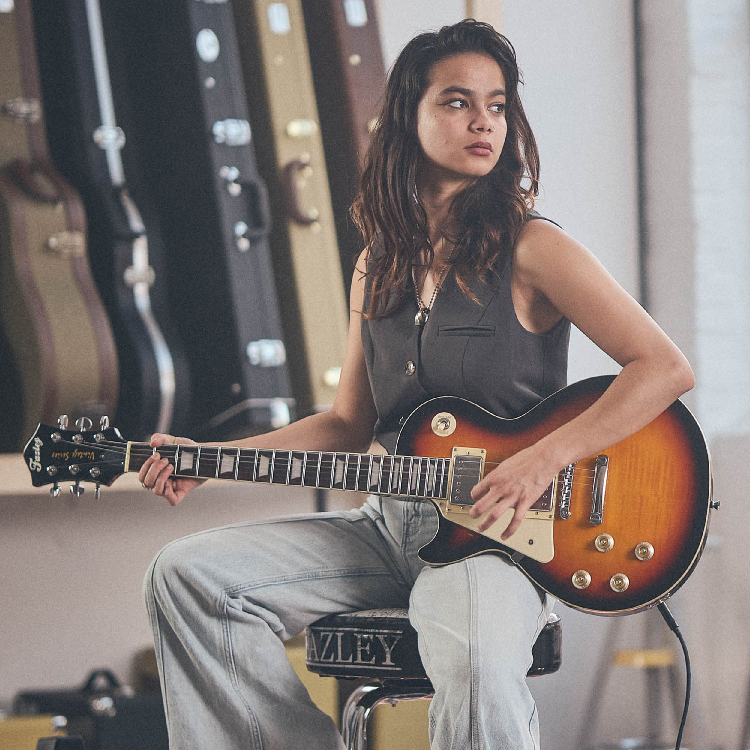 Een vrouw zit gitaar te spelen. Gitaarkoffers op de achtergrond.