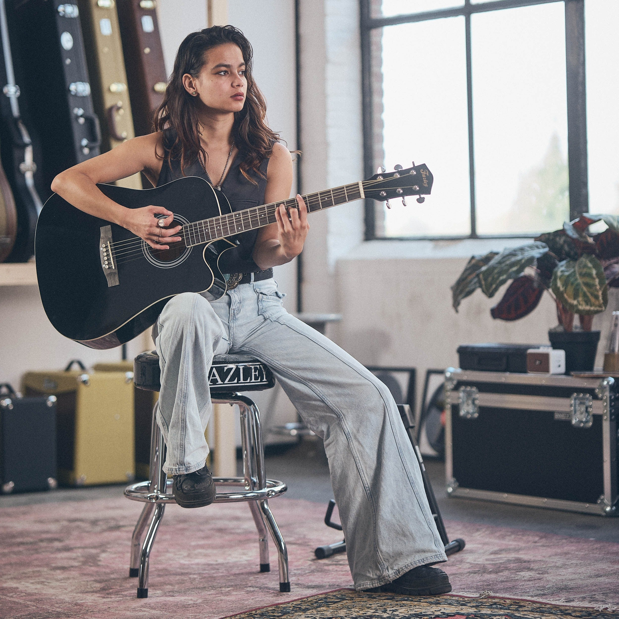 A woman sits on a stool playing a black guitar.
