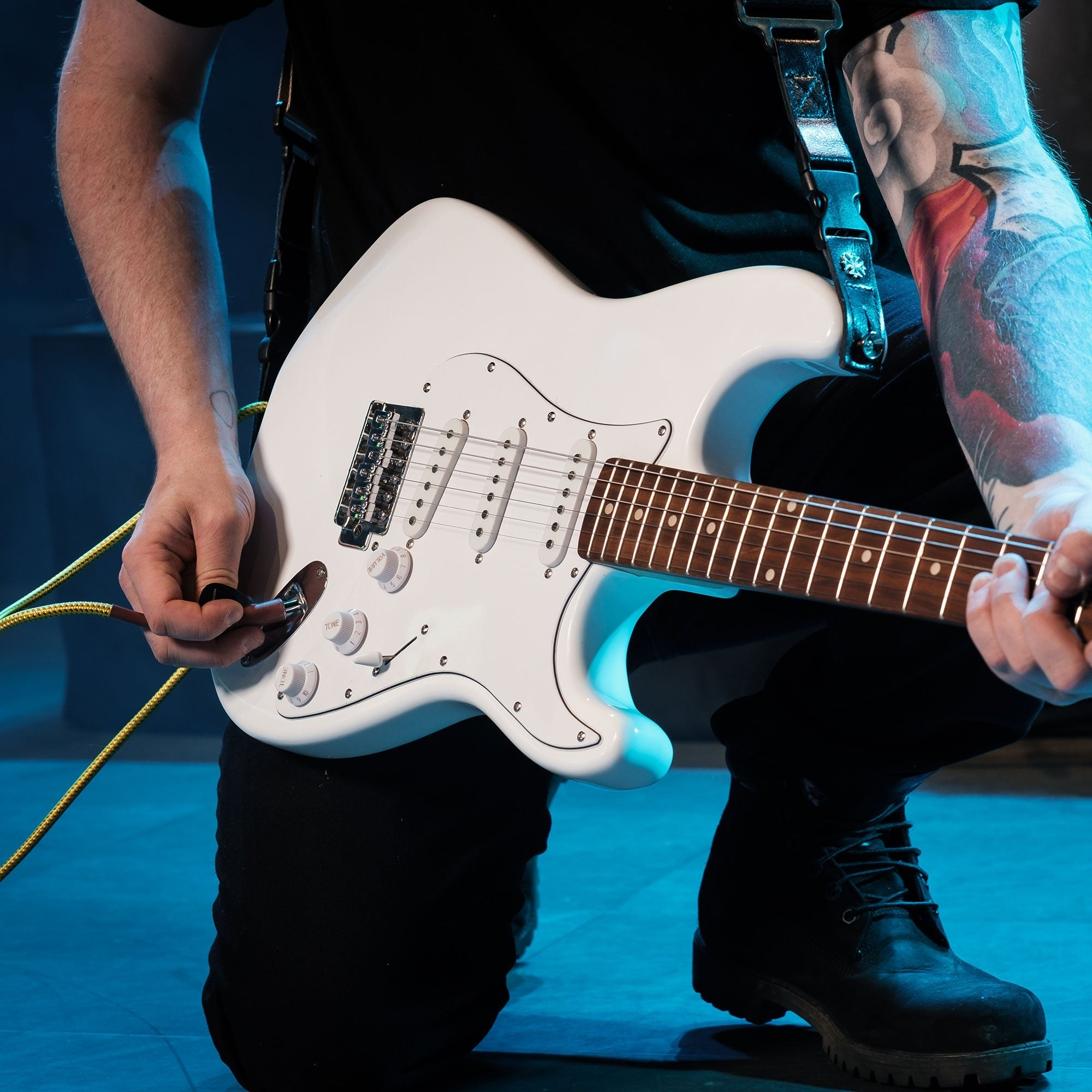 Persoon speelt witte elektrische gitaar, plugt kabel in. Blauwe verlichting.
