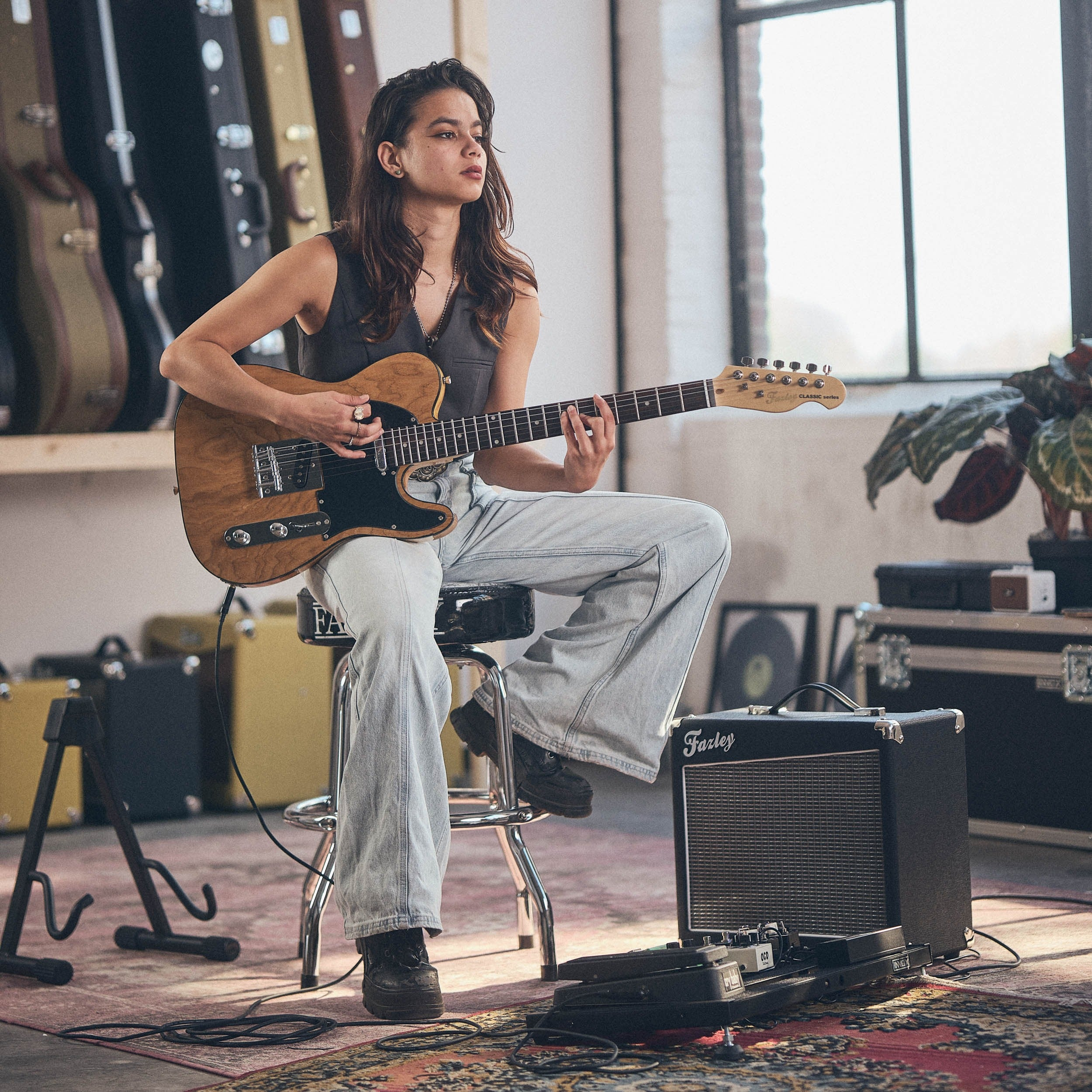 Jonge vrouw speelt gitaar op kruk, omringd door gitaarkoffers en versterkers.