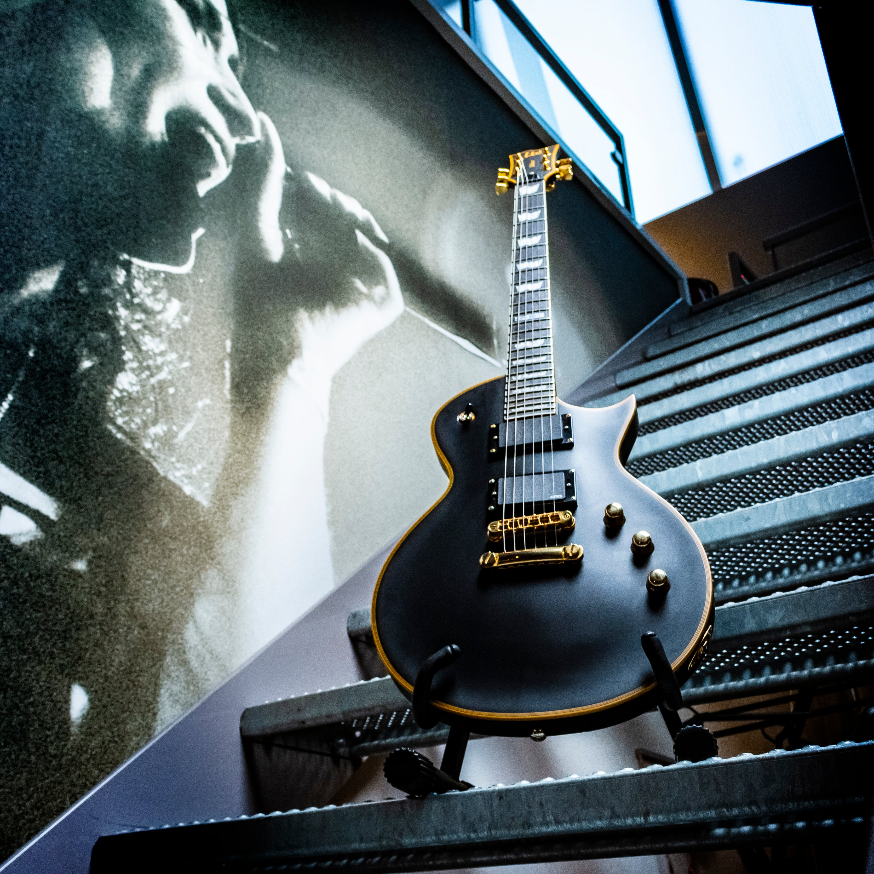 Black electric guitar with gold accents on metal stairs, black and white portrait in background.