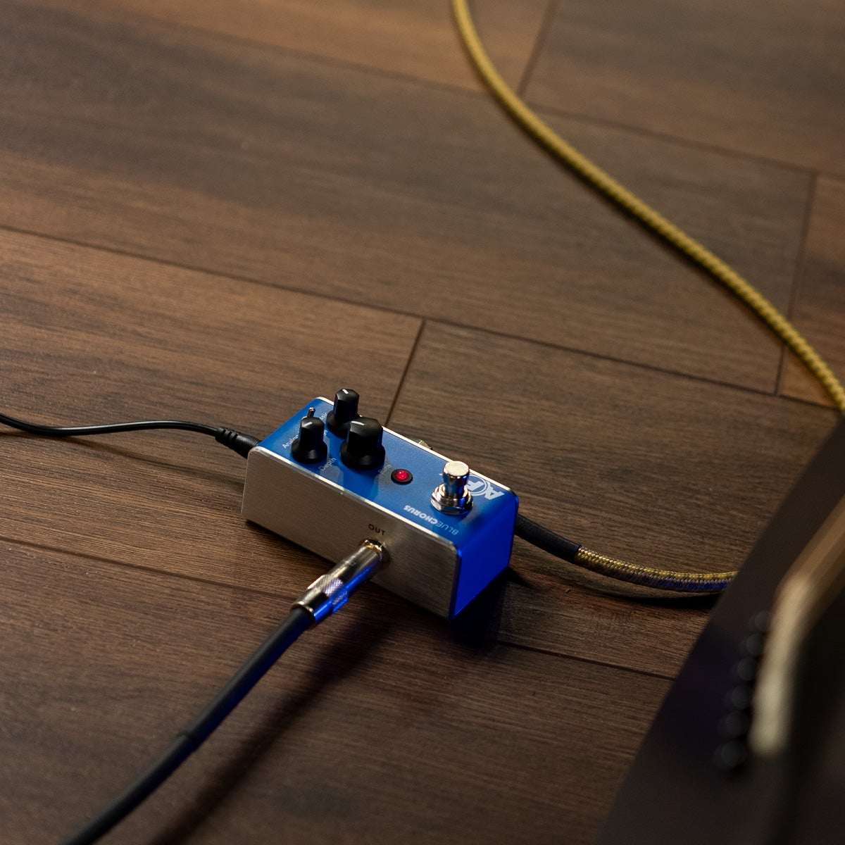Blue guitar effect pedal on wooden floor with cables.