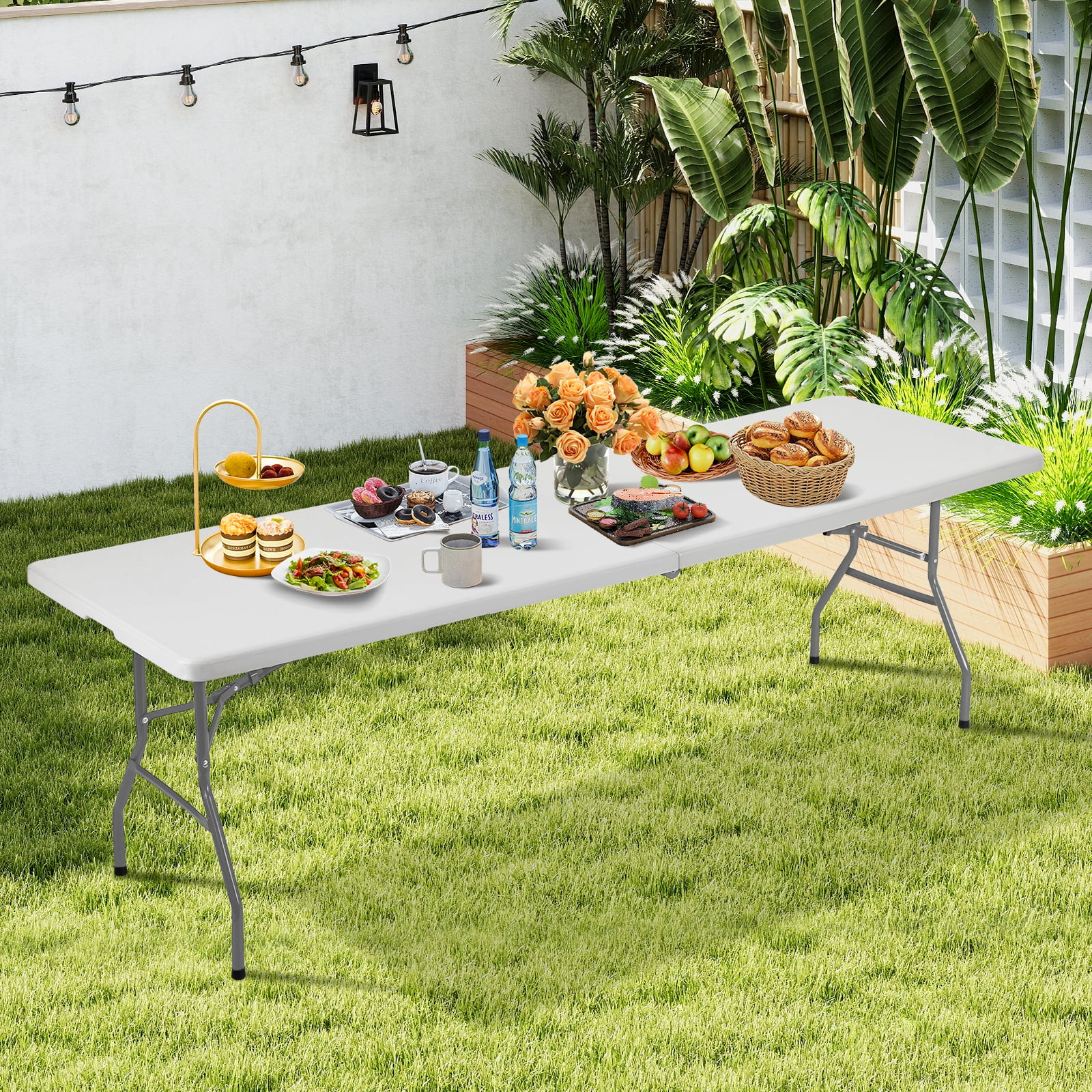Una mesa plegable blanca con comida sobre césped, plantas y luces de fondo.