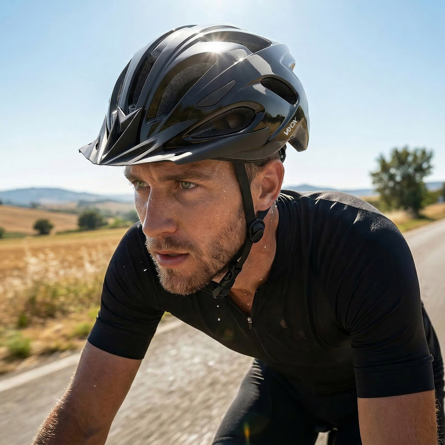 Mężczyzna w kasku jedzie rowerem po drodze, spocony, z wiejskim krajobrazem w tle.