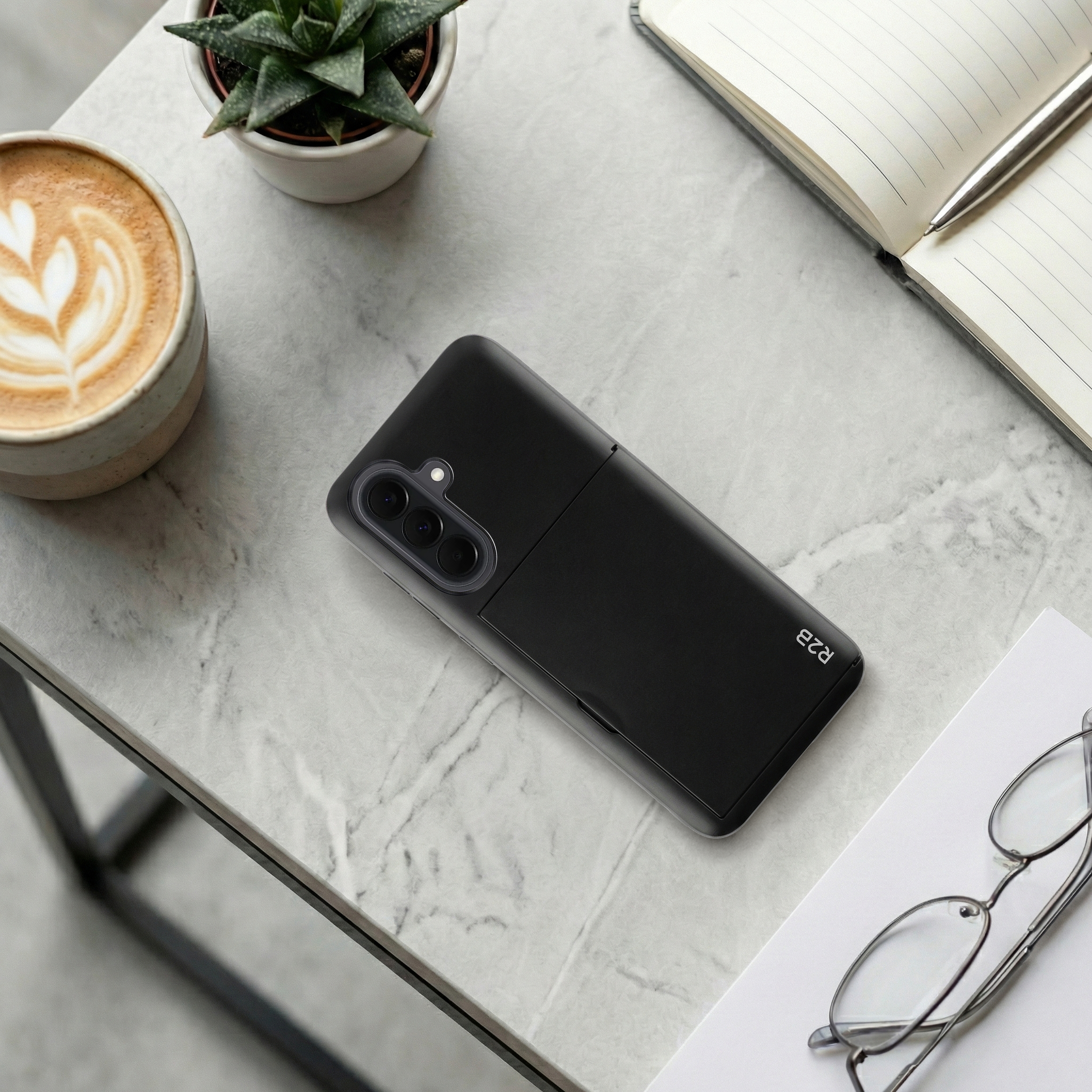 Schwarzes Smartphone auf Marmortisch mit Kaffee, Pflanze, Notizbuch und Brille.