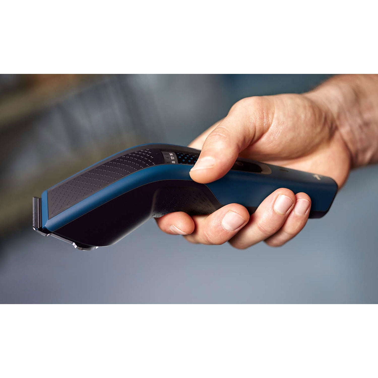 Een hand houdt een blauwe en zwarte tondeuse met metalen mesjes vast.