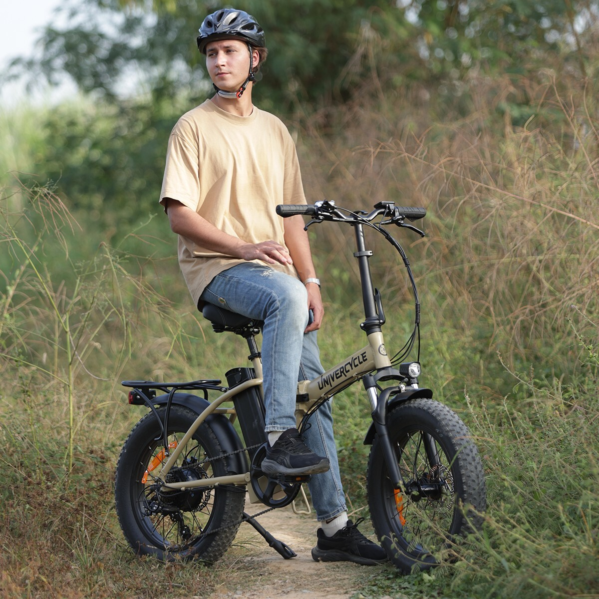 Mężczyzna w kasku siedzi na beżowym rowerze elektrycznym na zewnątrz, otoczony zielenią.
