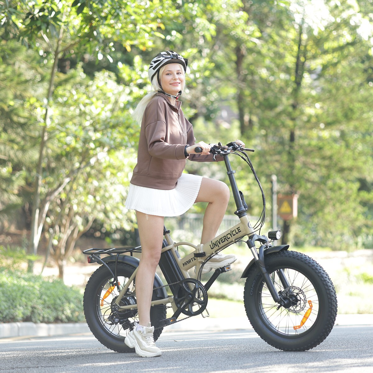 Kobieta w brązowym swetrze i białej spódnicy jedzie na beżowym rowerze elektrycznym na zewnątrz.
