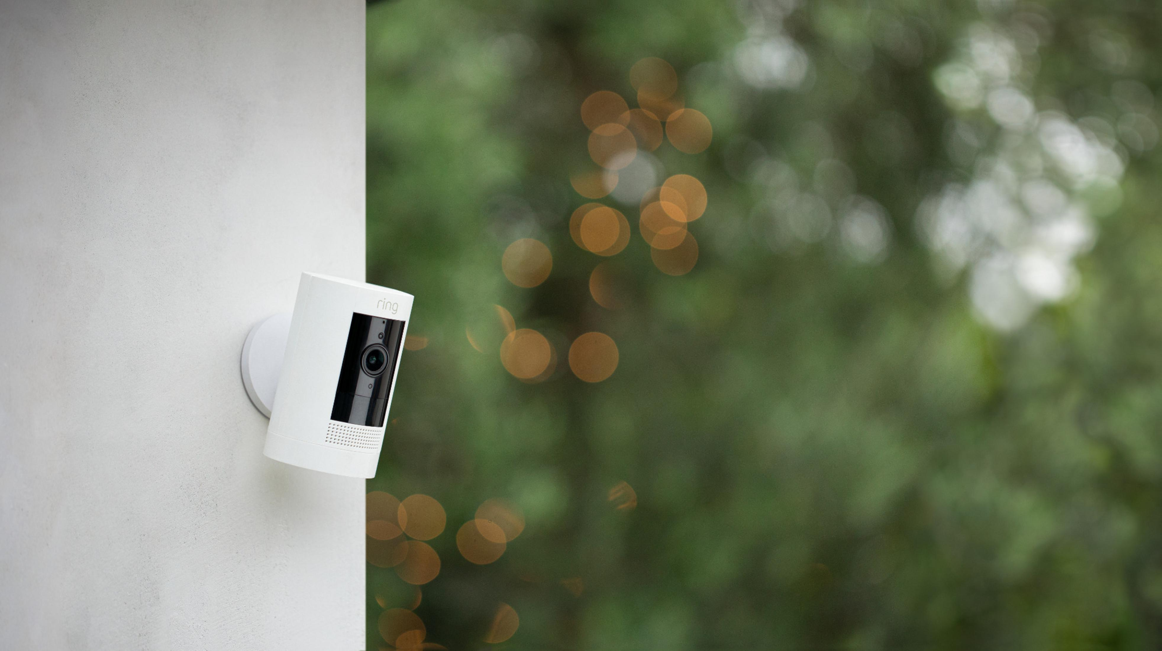 Biała kamera Ring zamontowana na ścianie. W tle drzewa.