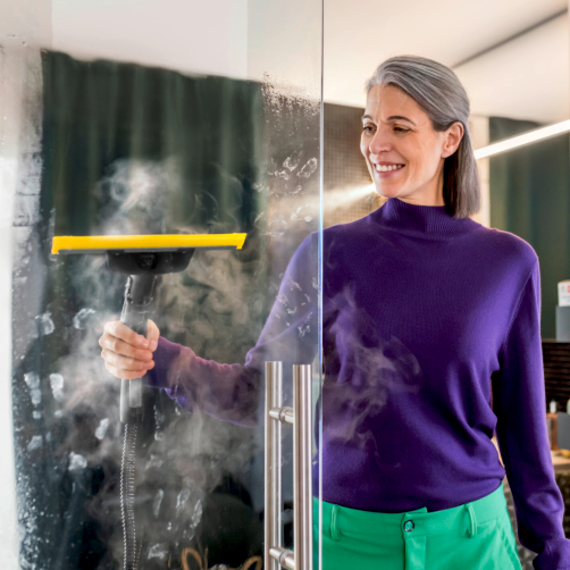 Donna con top viola e pantaloni verdi pulisce a vapore una porta di vetro.