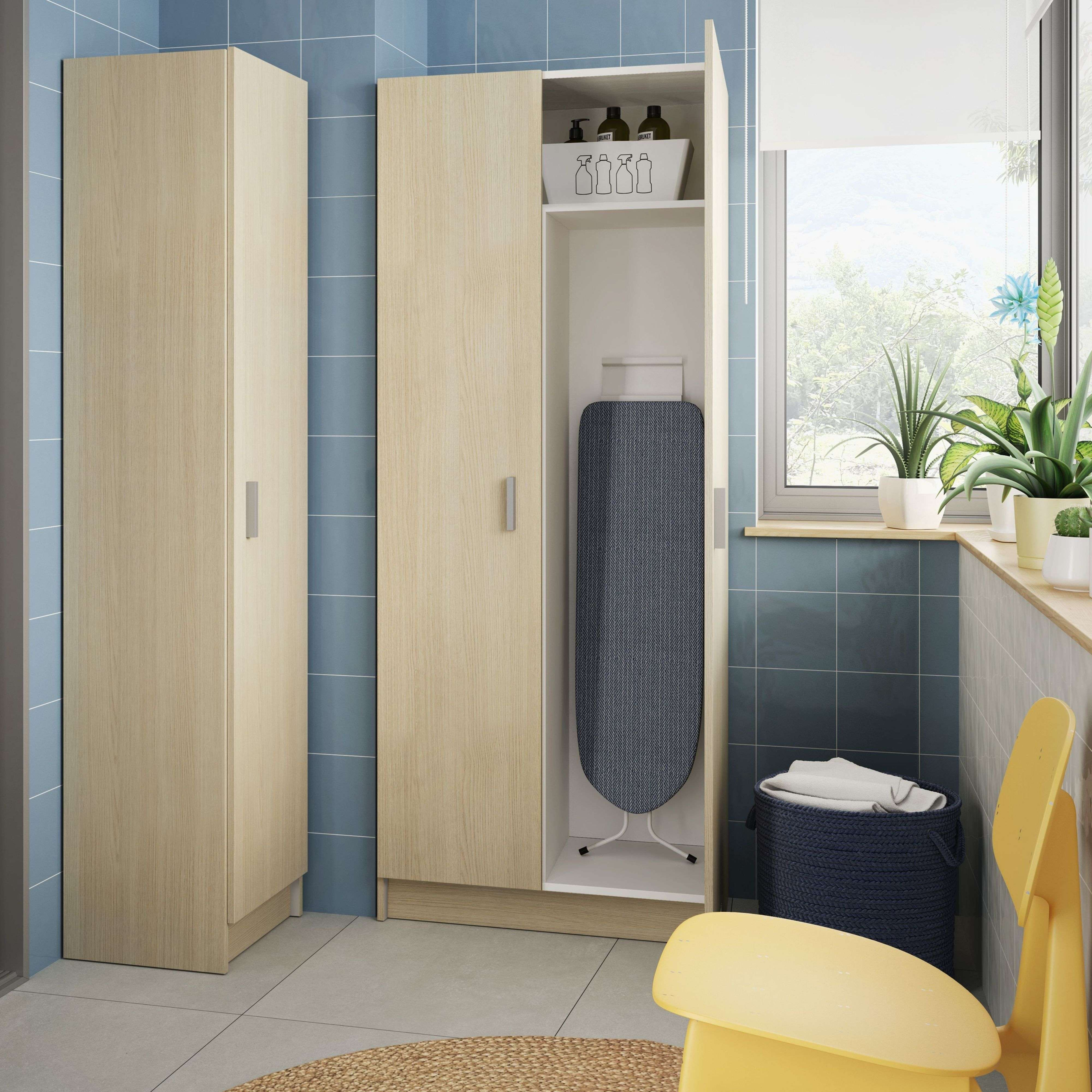 Armarios de madera en baño con tabla de planchar, cesto y plantas junto a ventana.