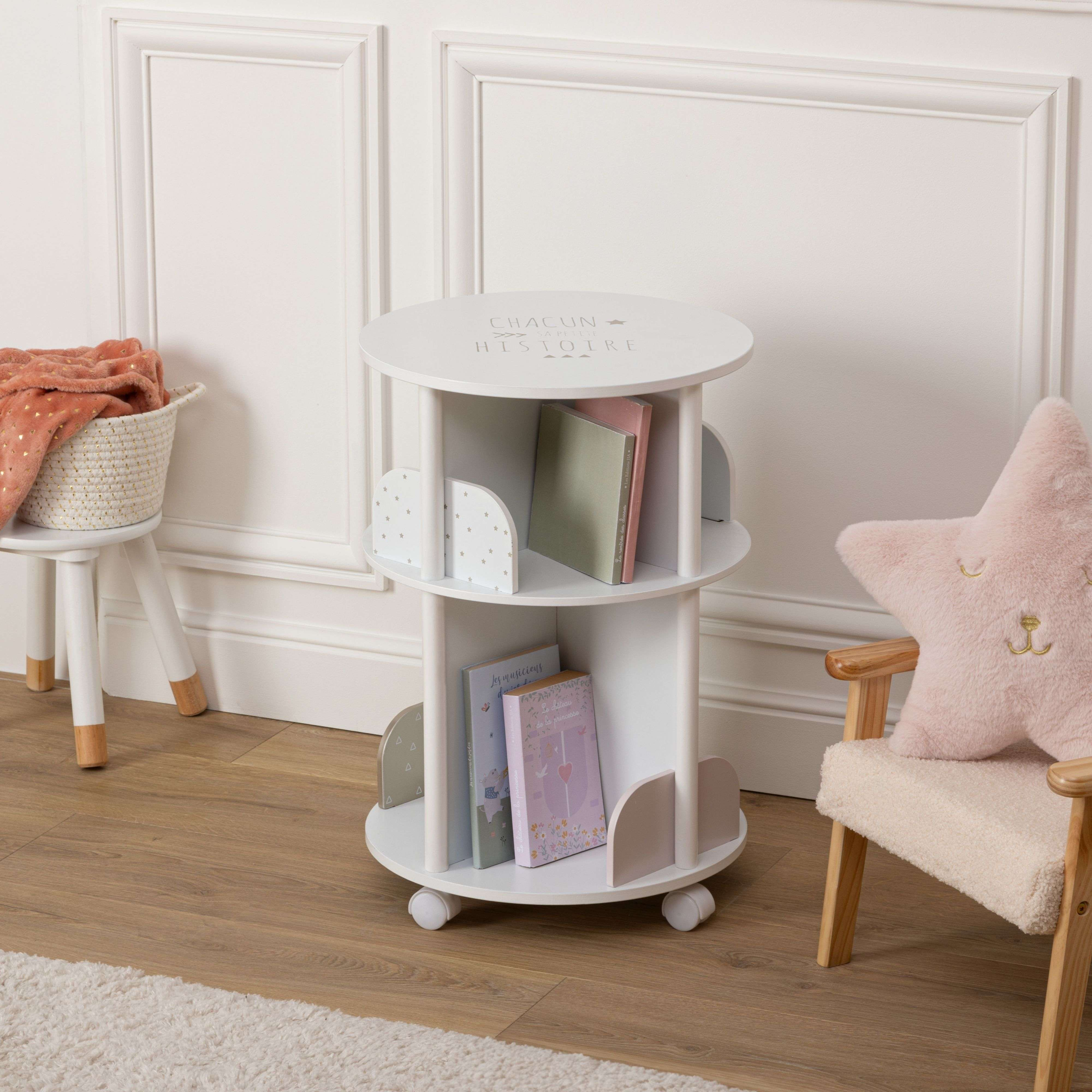Un librero blanco con libros, al lado una silla con cojín de estrella y una cesta con manta.