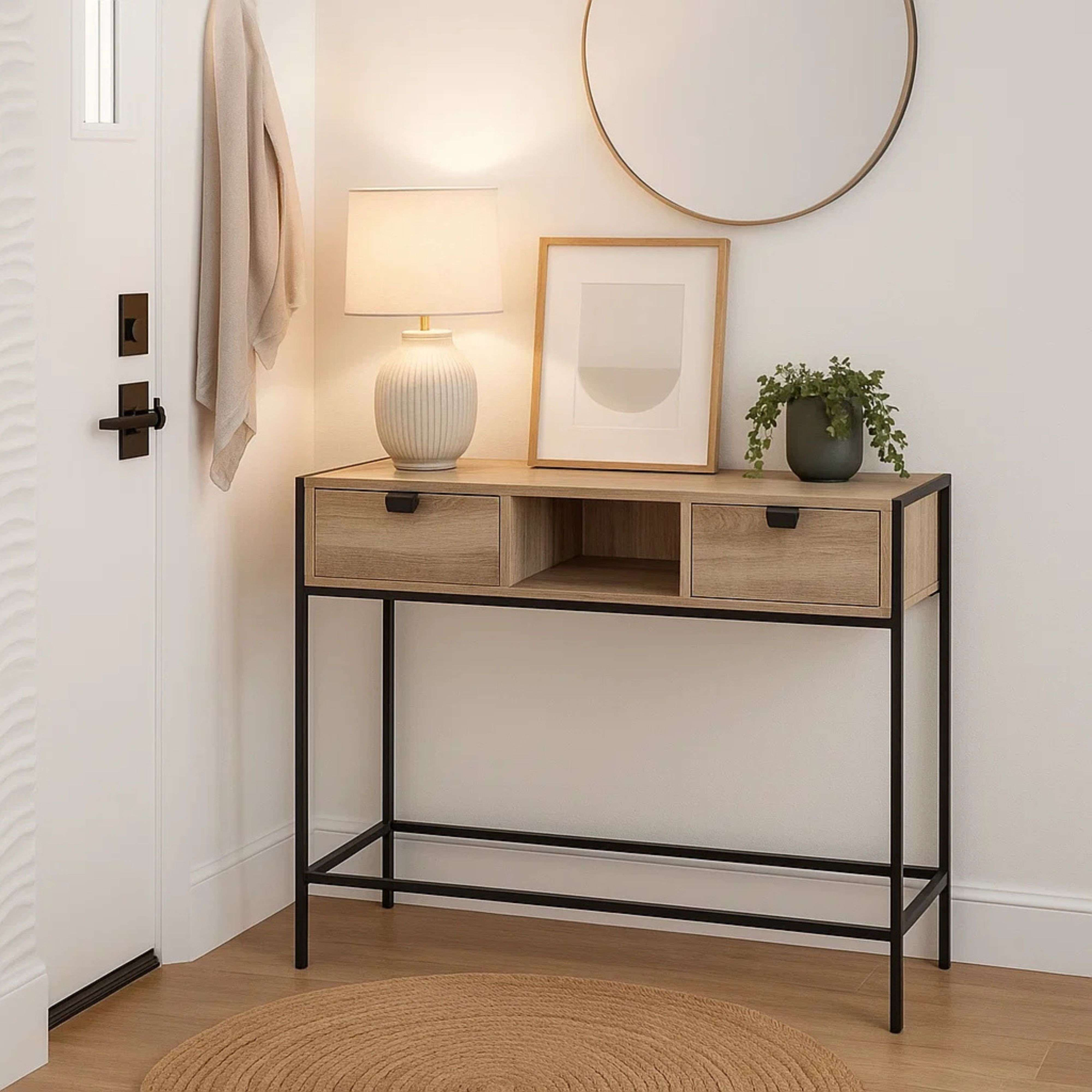 Una consola estrecha de madera con patas negras, lámpara, cuadro y planta.