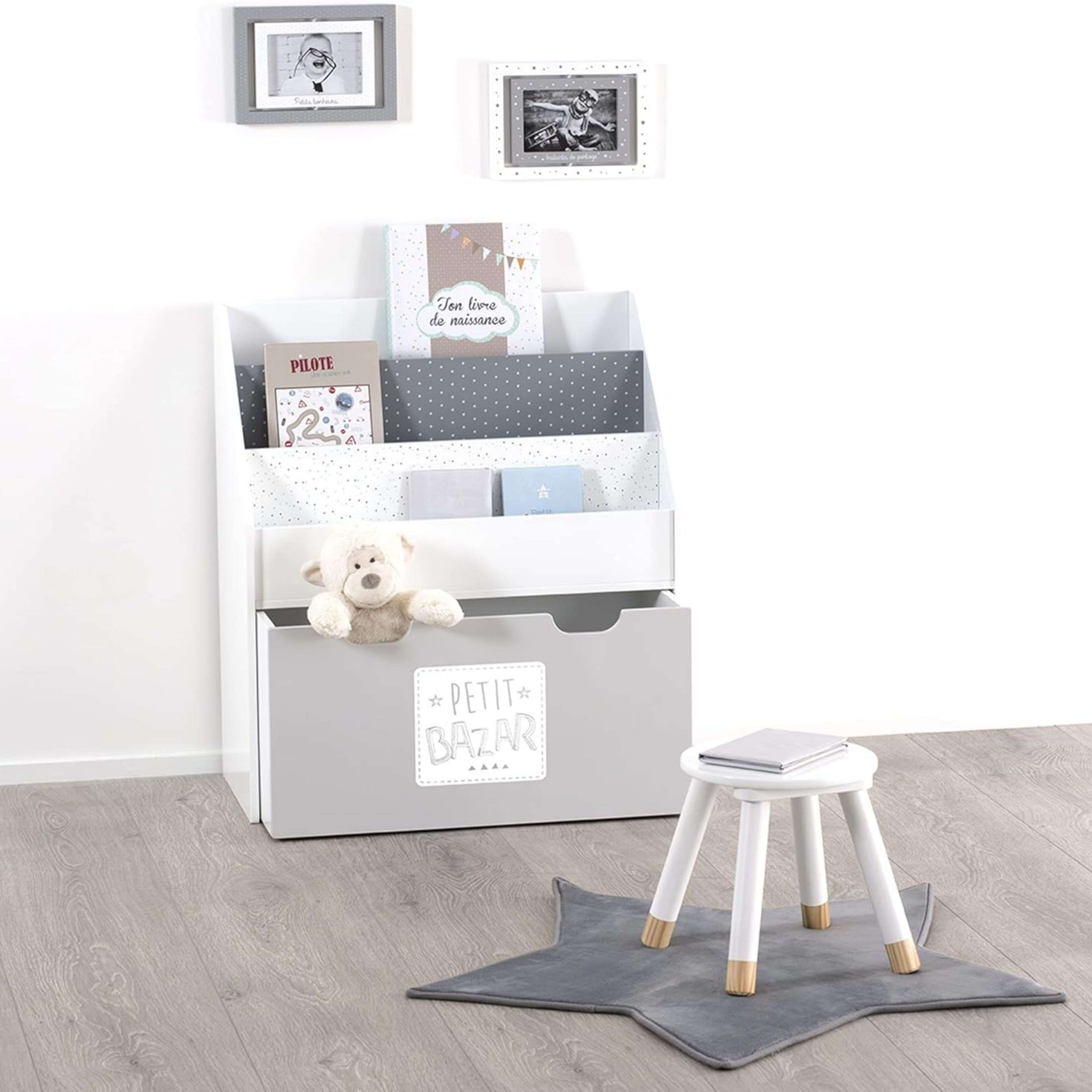 Estantería blanca con libros, osito y caja. Alfombra estrella y taburete cerca.