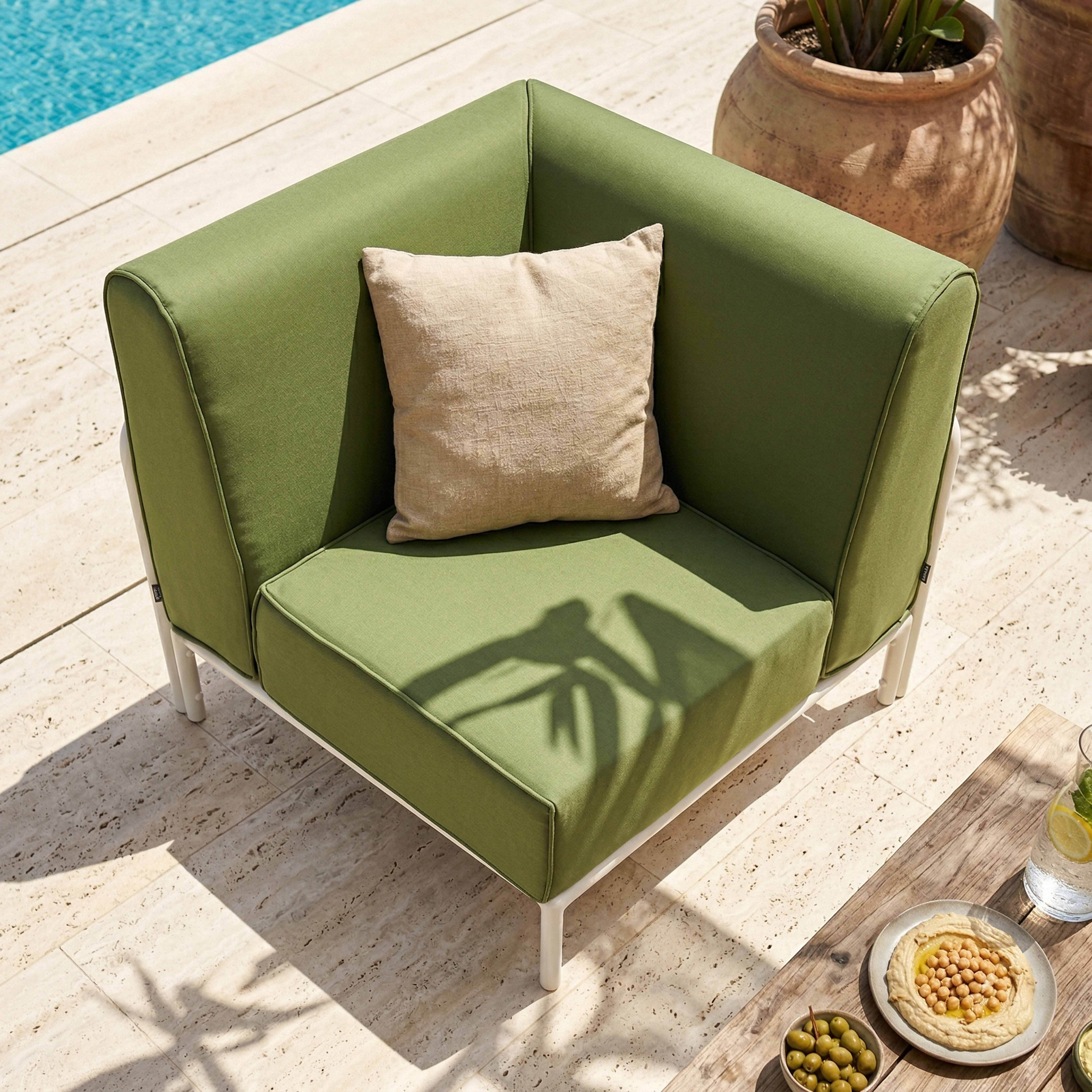 Grünes Ecksofa mit Beigekissen am Pool. Hummus und Oliven auf Holztisch.