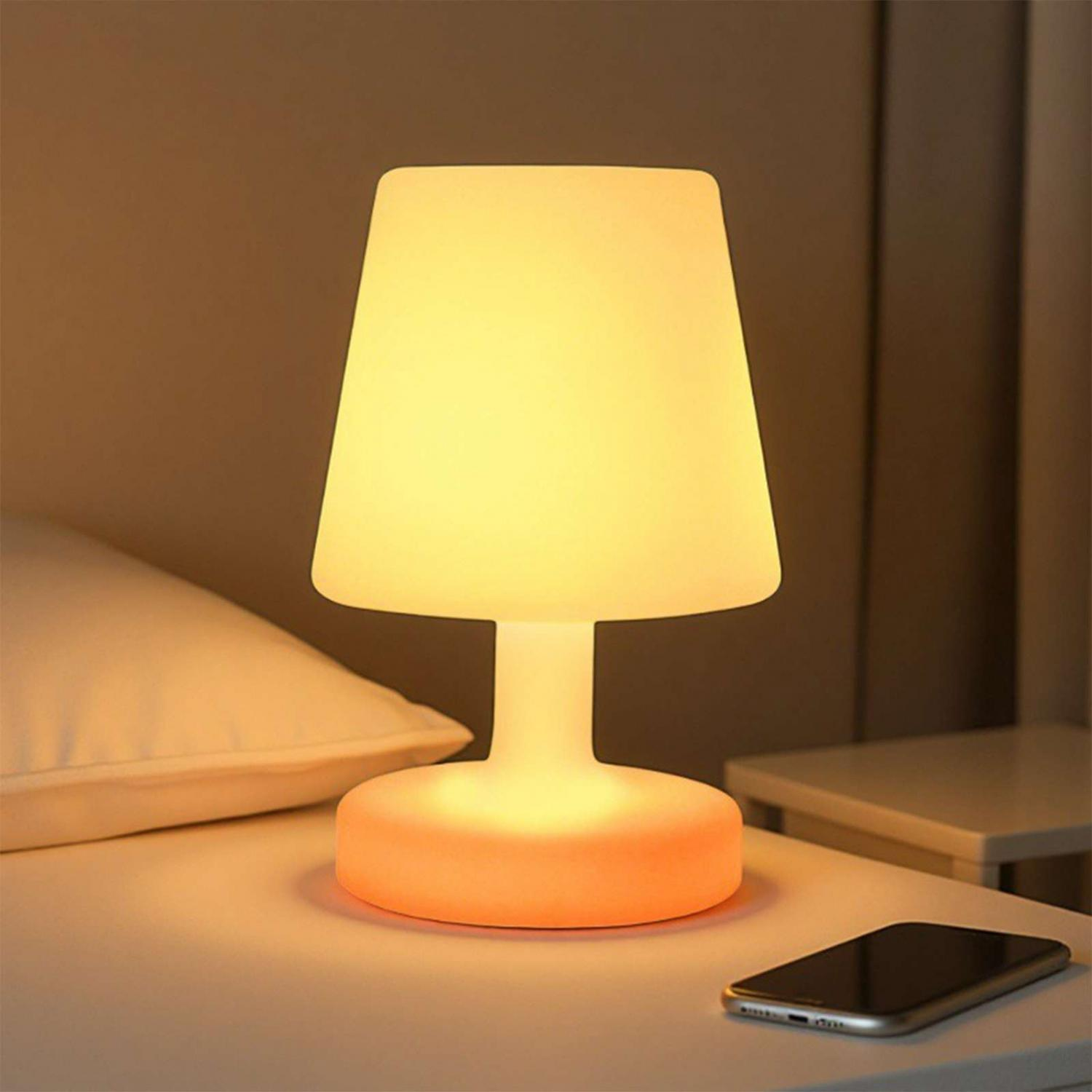 Eine weiße Nachttischlampe leuchtet warm gelb und orange neben Kissen und Handy.