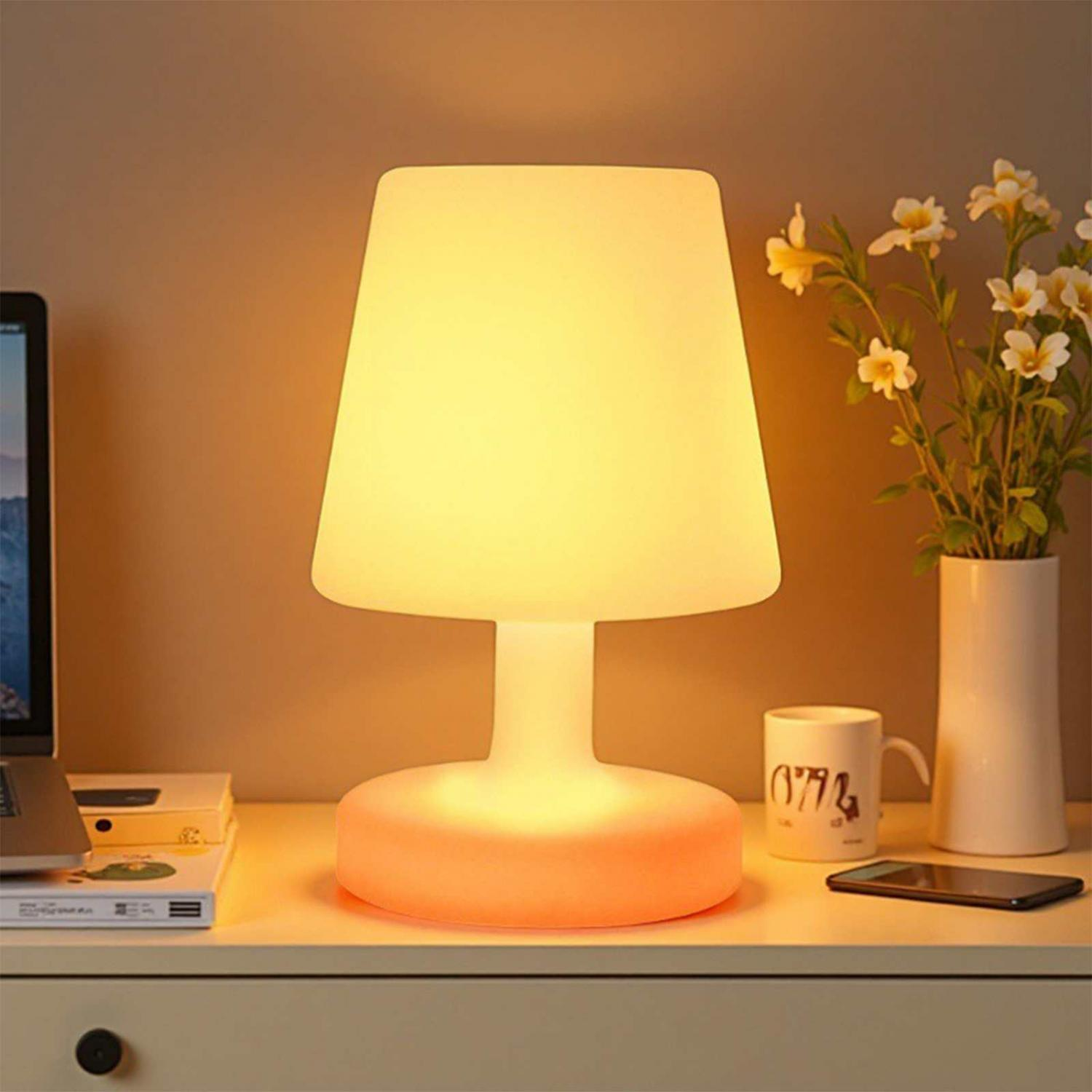 Eine leuchtende weiße Lampe steht auf einem Schreibtisch mit Laptop, Büchern, Tasse, Handy und Blumen.