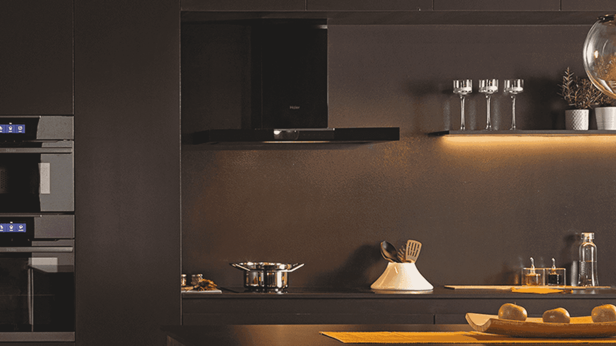 Moderna cocina oscura con campana extractora, hornos, vasos y frutero.