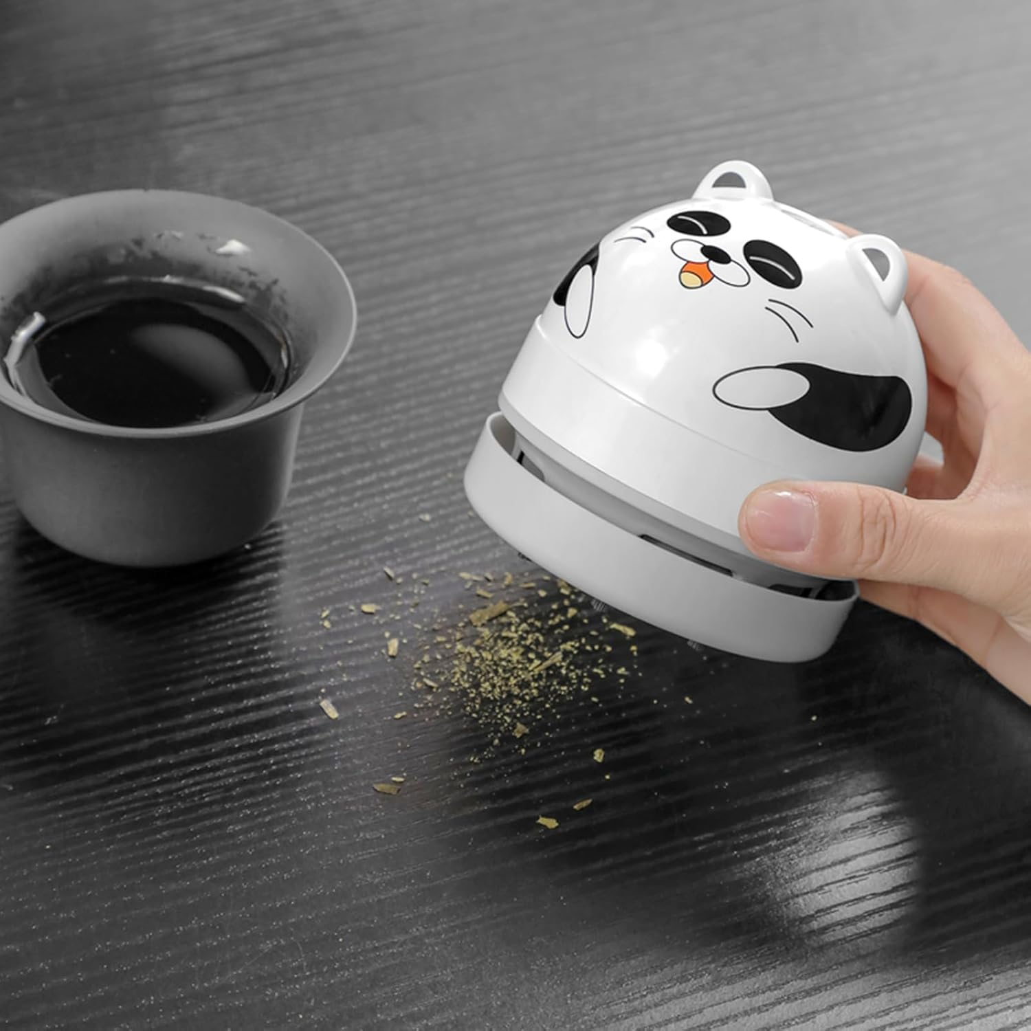 Eine Hand hält einen weißen Mini-Staubsauger in Panda-Form, der Krümel auf einem Holztisch aufsaugt.