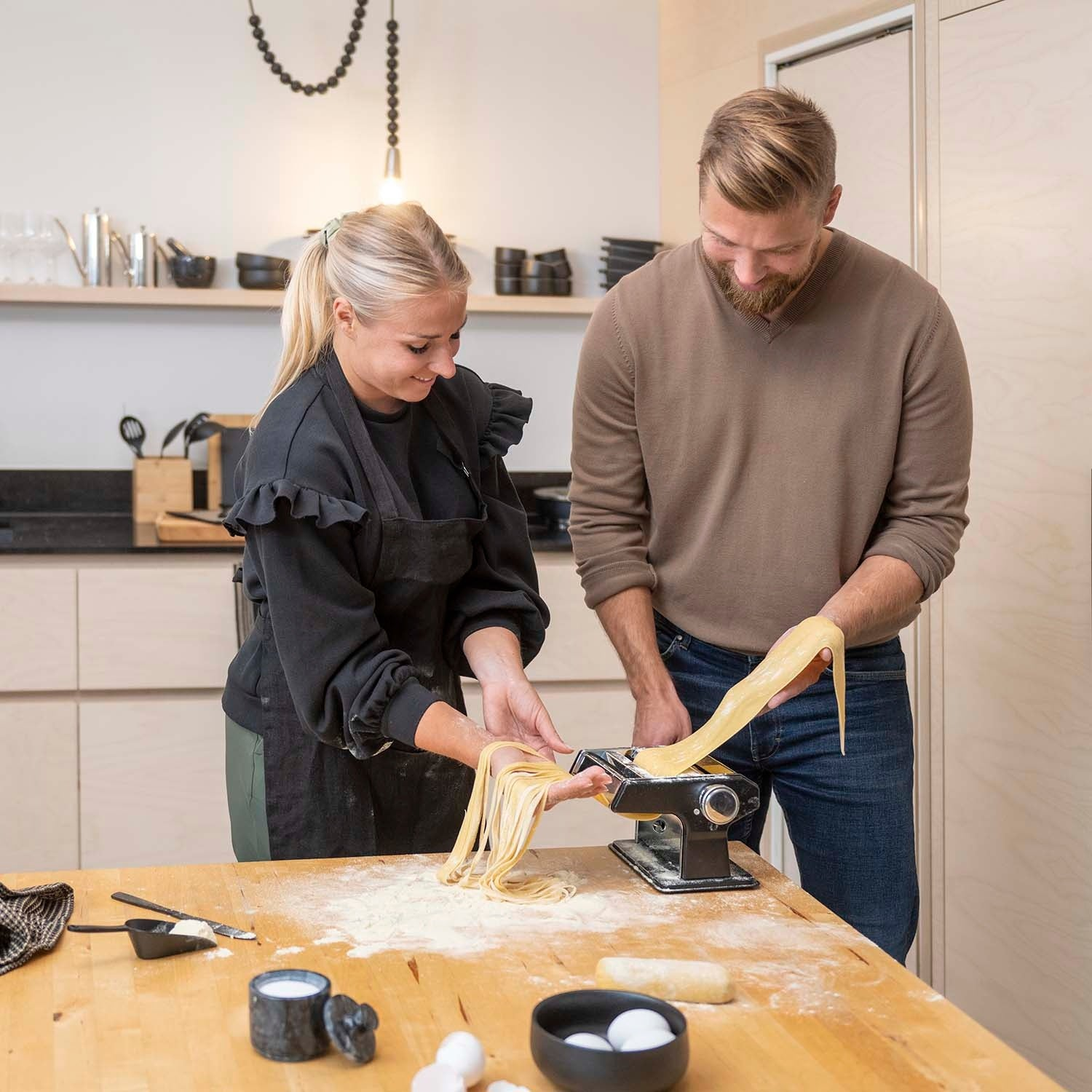 A man and woman make pasta using a machine in a modern kitchen. Flour is on the table.