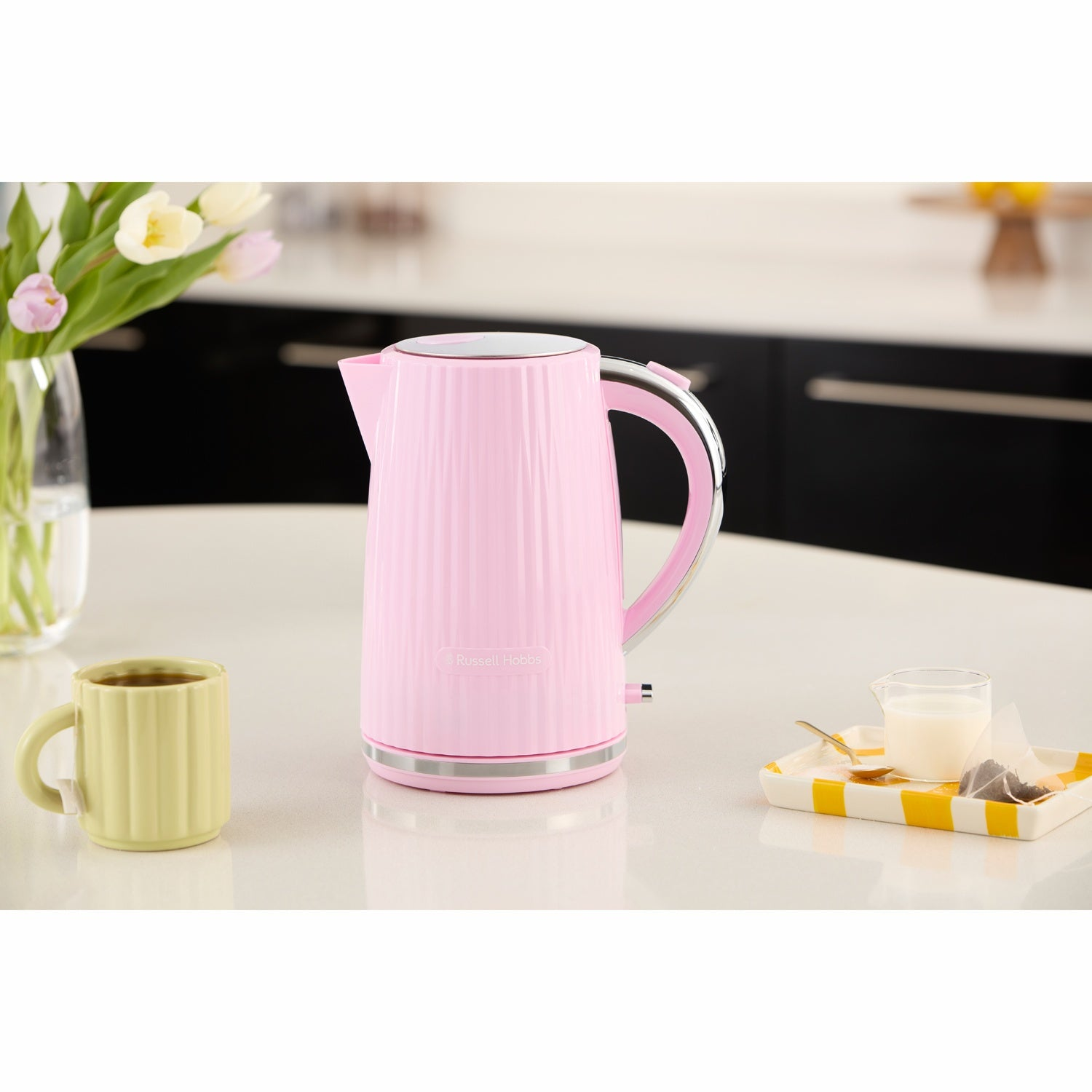 Een roze waterkoker staat op een aanrecht naast een mok koffie en bloemen.