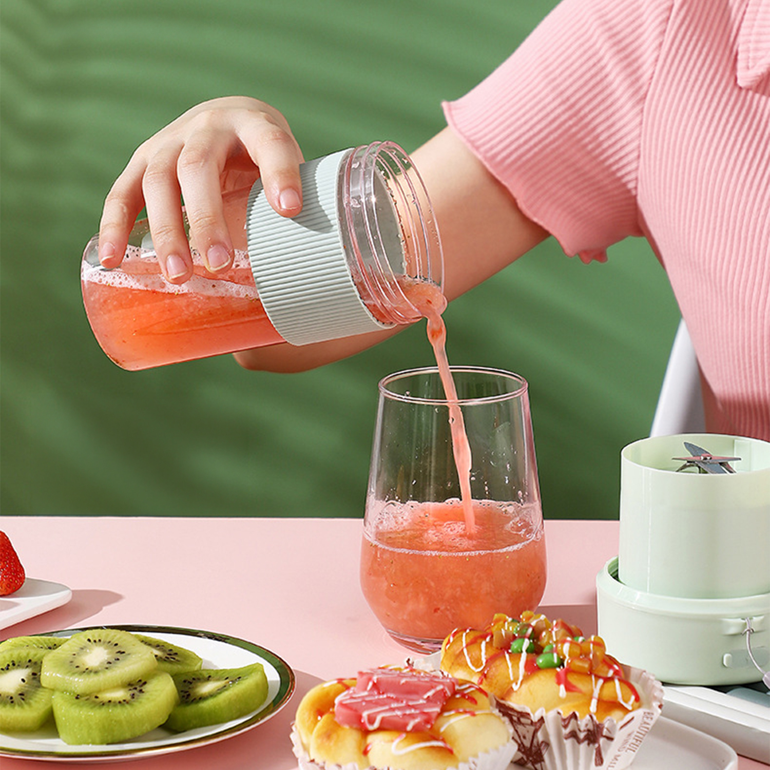 Una mano vierte zumo rosa de una licuadora en un vaso. Kiwi, fresa y pasteles están sobre la mesa.