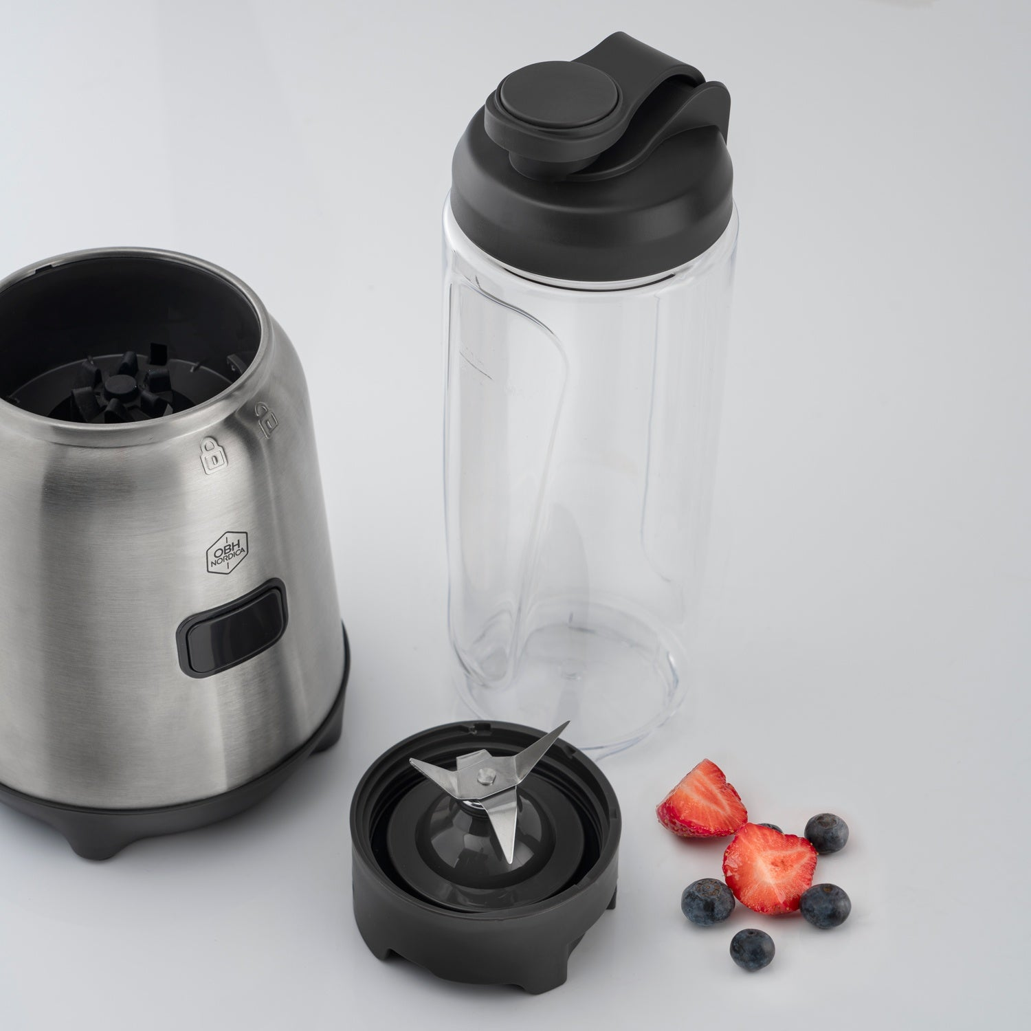 A silver blender base, blade set, and clear container with lid. Strawberries and blueberries are nearby.