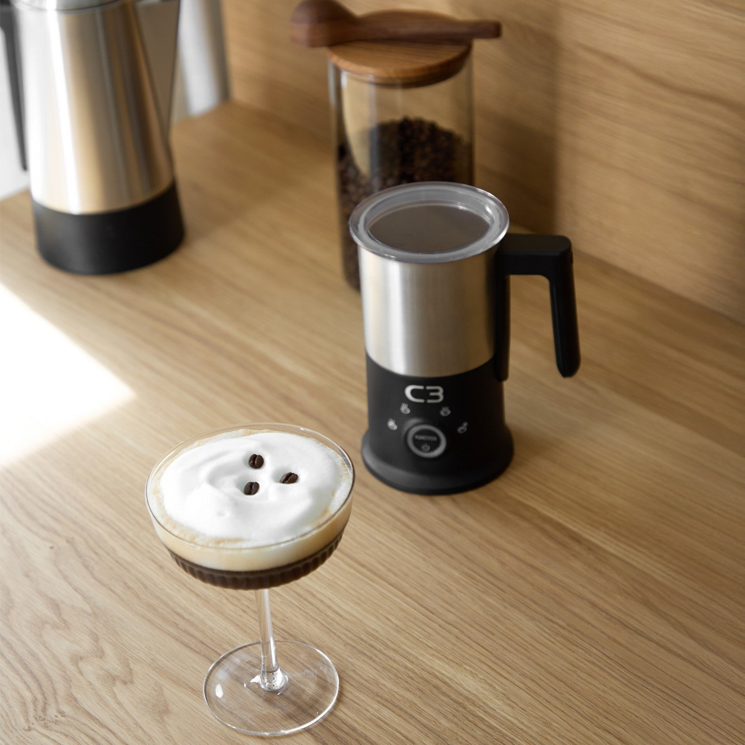 A glass with a layered coffee drink sits on a wooden table beside a milk frother and coffee beans.