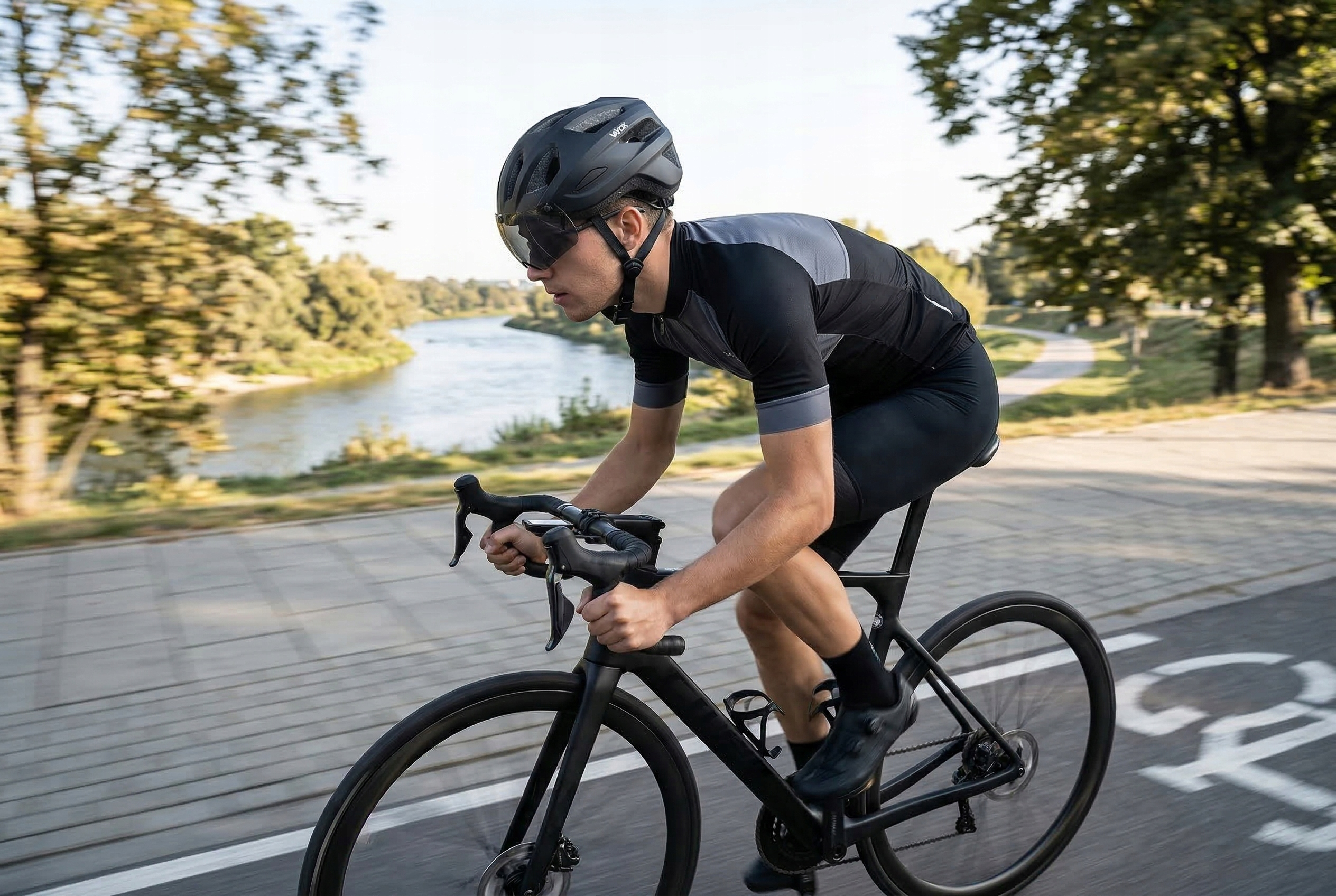 Mężczyzna jedzie rowerem po ścieżce nad rzeką, w kasku i stroju kolarskim.