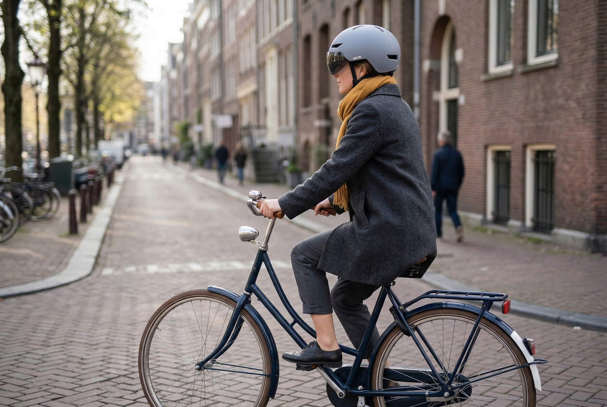 Kobieta jedzie rowerem po ulicy. Ma na sobie szary płaszcz, żółty szalik i kask.