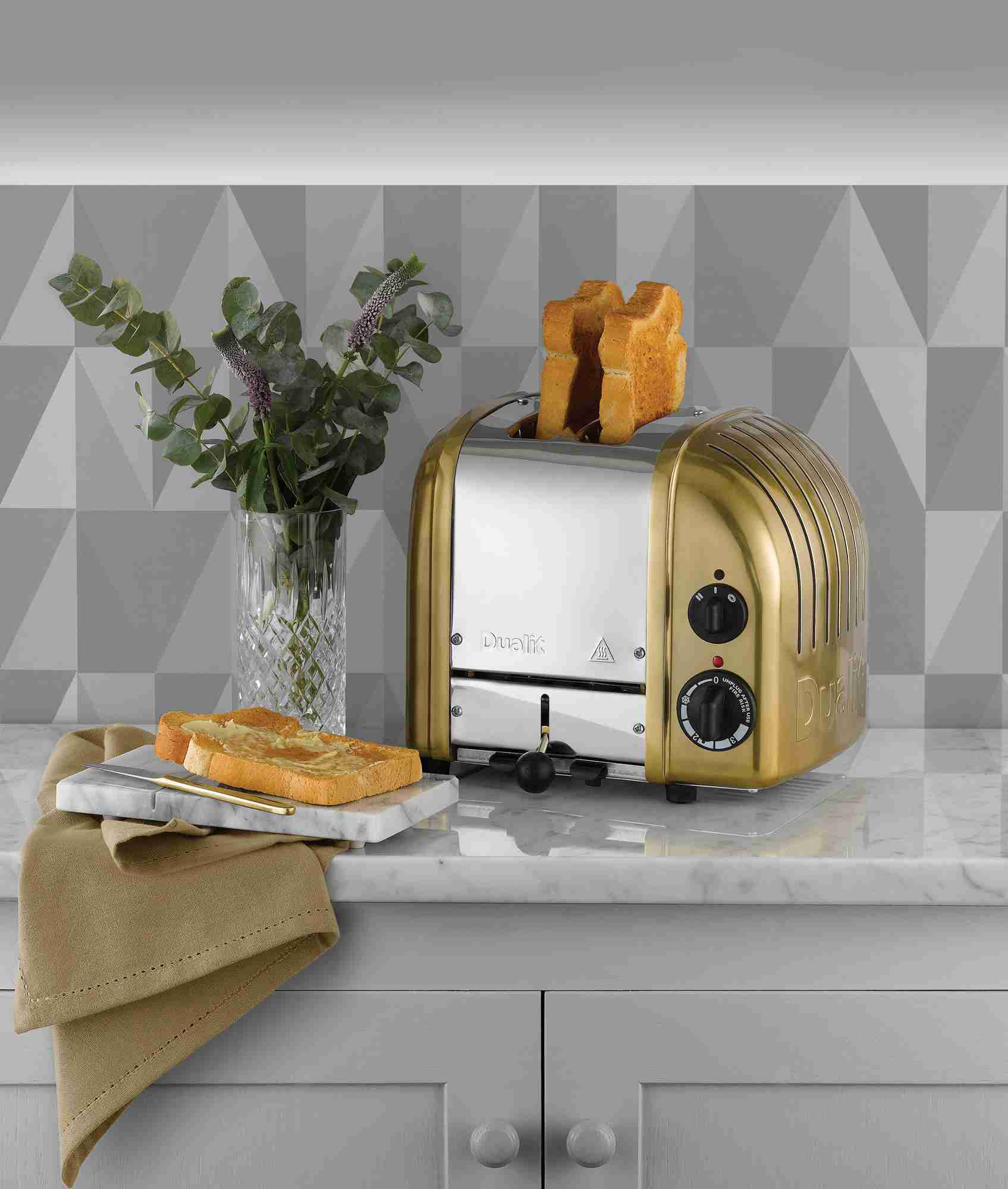 Ein goldener Toaster mit zwei Toastbroten, daneben ein Glas mit Blumen und Buttertoast.