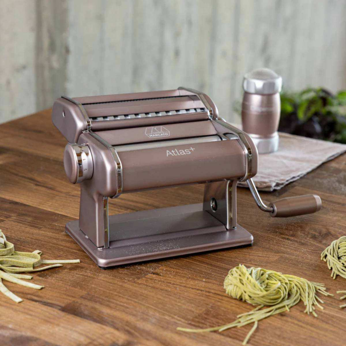Eine bronzefarbene Nudelmaschine mit Handkurbel steht auf einem Holztisch mit Nudeln.